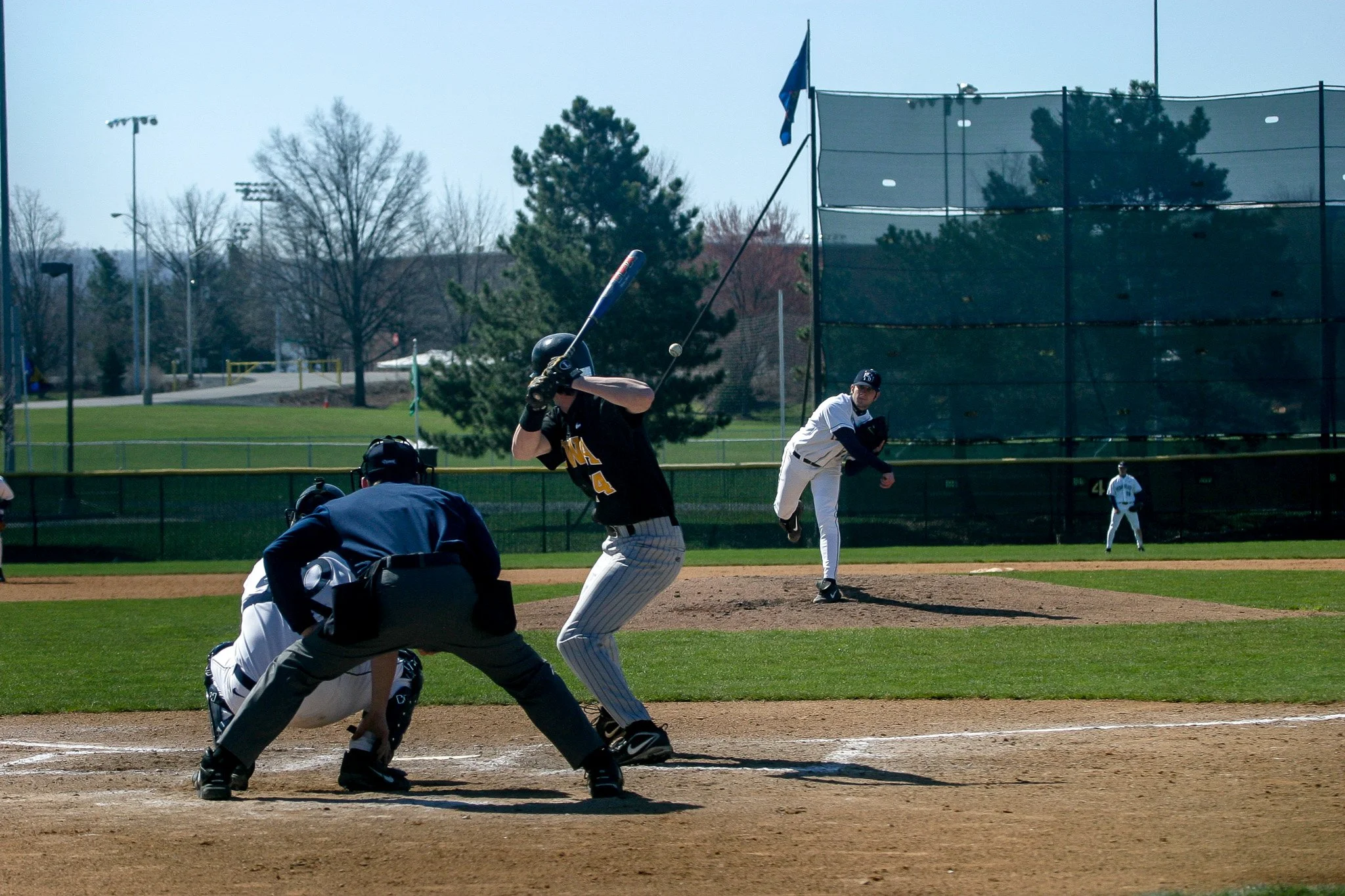 A baseball game in progress with a batter swinging at a pitch, a catcher crouched behind home plate, a pitcher throwing, and an umpire behind the catcher. Other players are visible in the outfield.