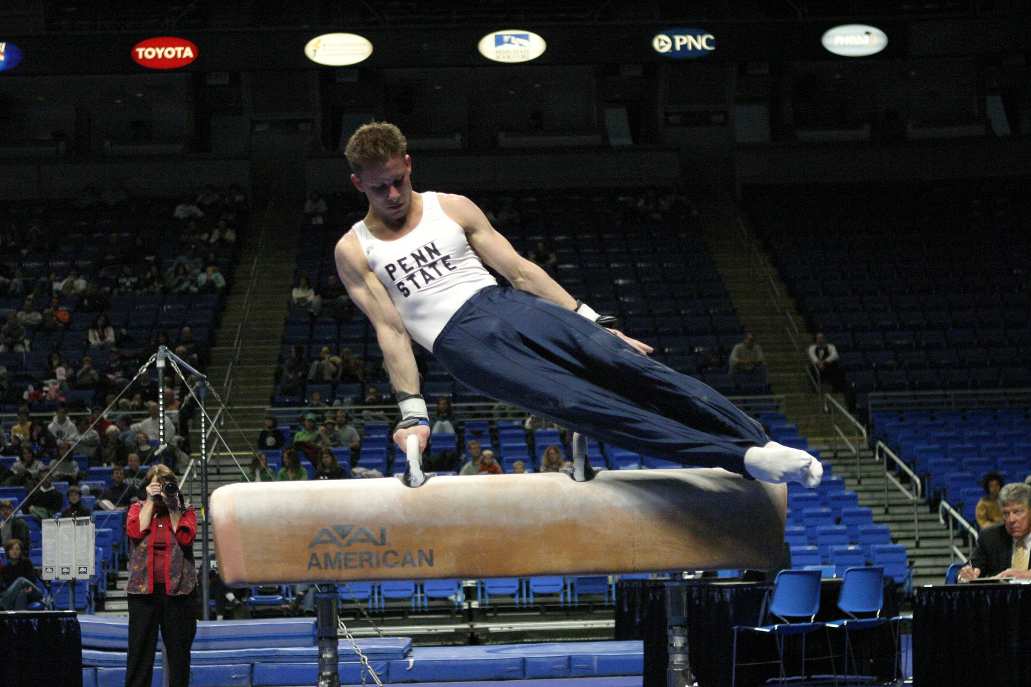 A male gymnast from Penn State performing a routine on the pommel horse during a gymnastics competition in an indoor arena filled with spectators.