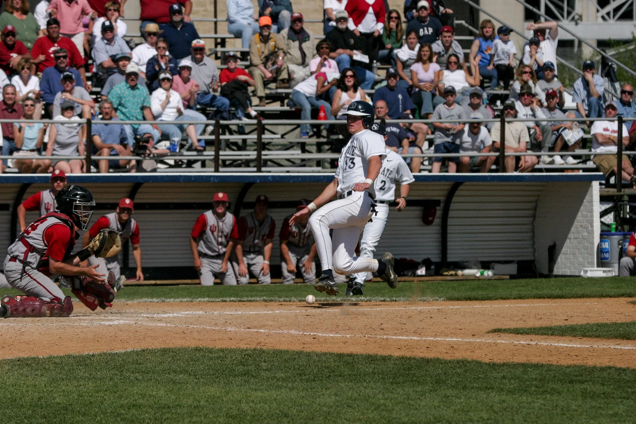 A baseball player in a white uniform is jumping to catch a baseball near home plate, with a catcher in red gear crouched in front. The crowd is watching from the bleachers.