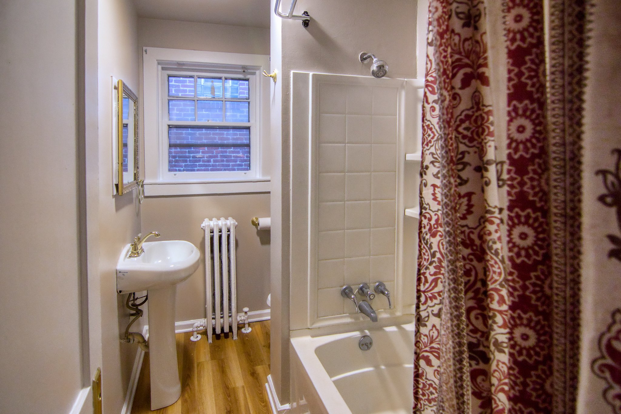 Small bathroom with beige walls, a window showing a brick building outside, a white pedestal sink, a white radiator, a bathtub with a showerhead, and a floral shower curtain.