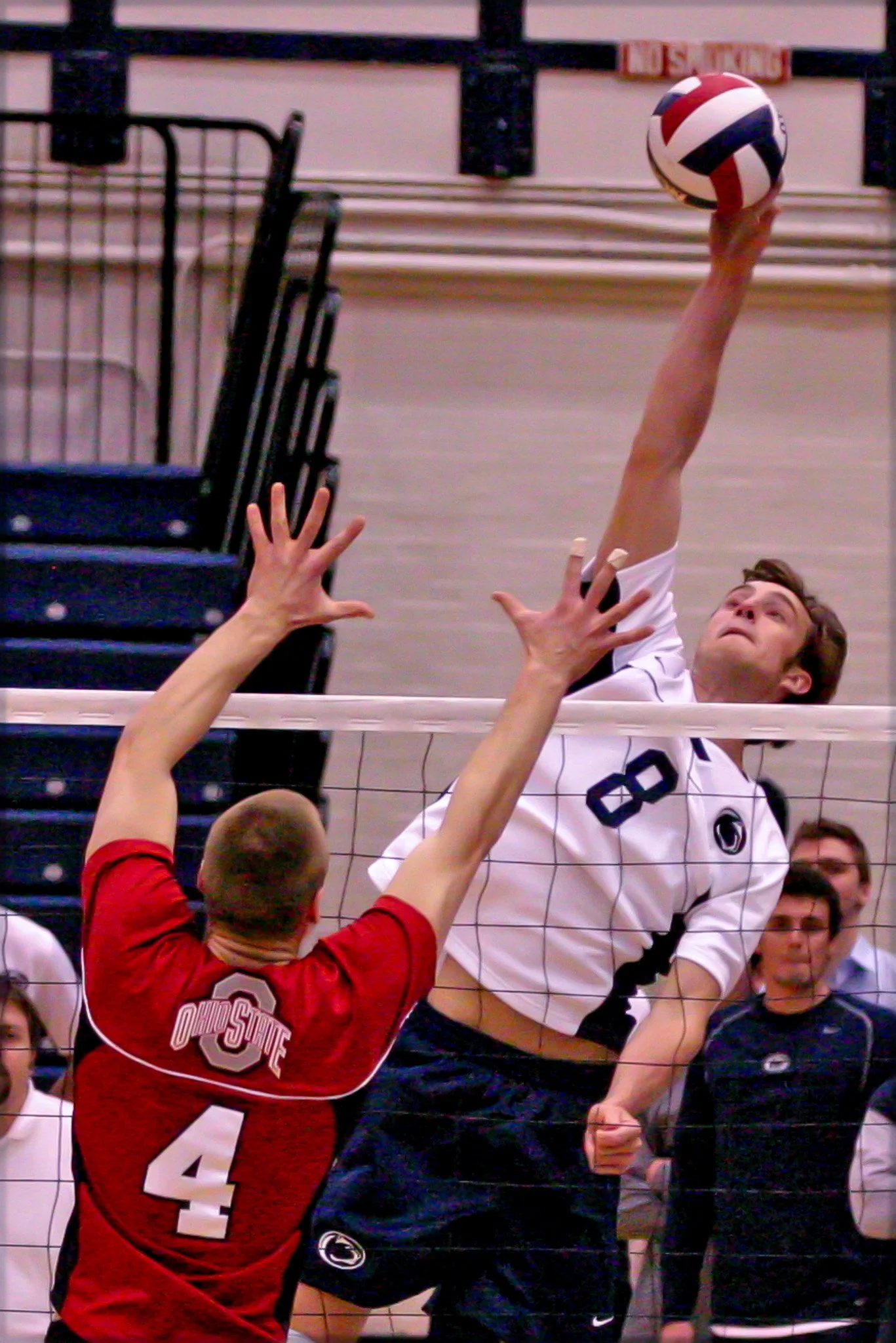 A volleyball player in a white Penn State uniform spikes the ball over the net while a player in a red Ohio State jersey attempts to block.