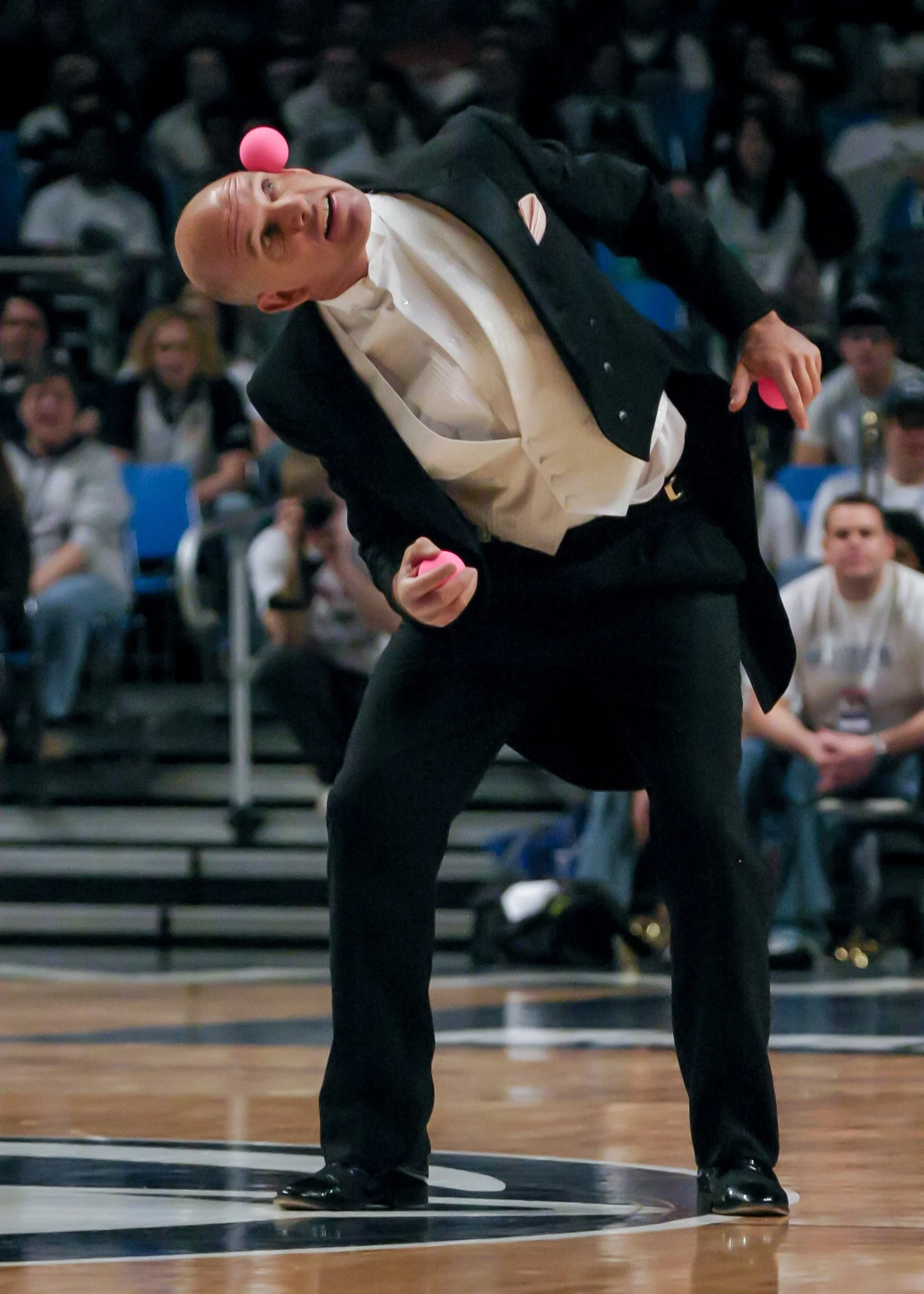 A man with a bald head dressed in a tuxedo, juggling three pink balls in an indoor arena with an audience watching.