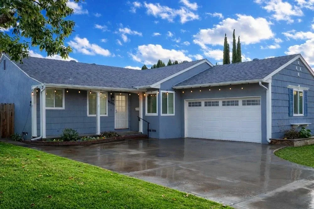 A blue single-story house with white trim, a driveway, and string lights hanging outside, under a partly cloudy sky.