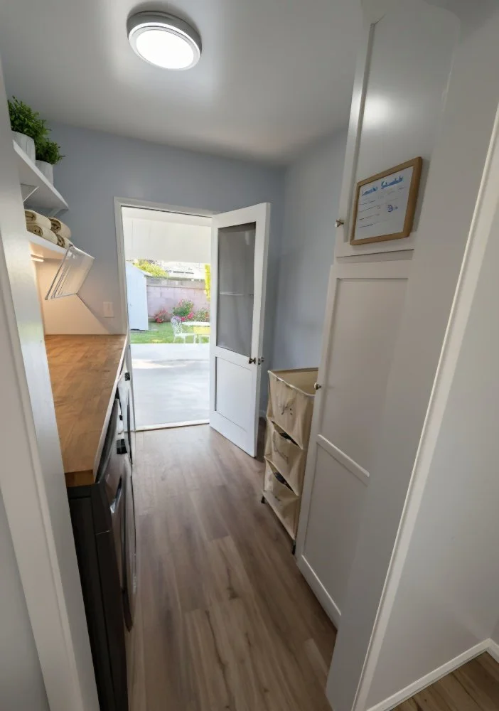 A small laundry room with a washer and dryer, a door leading outside, a wooden countertop, and shelves with folded towels and potted plants.