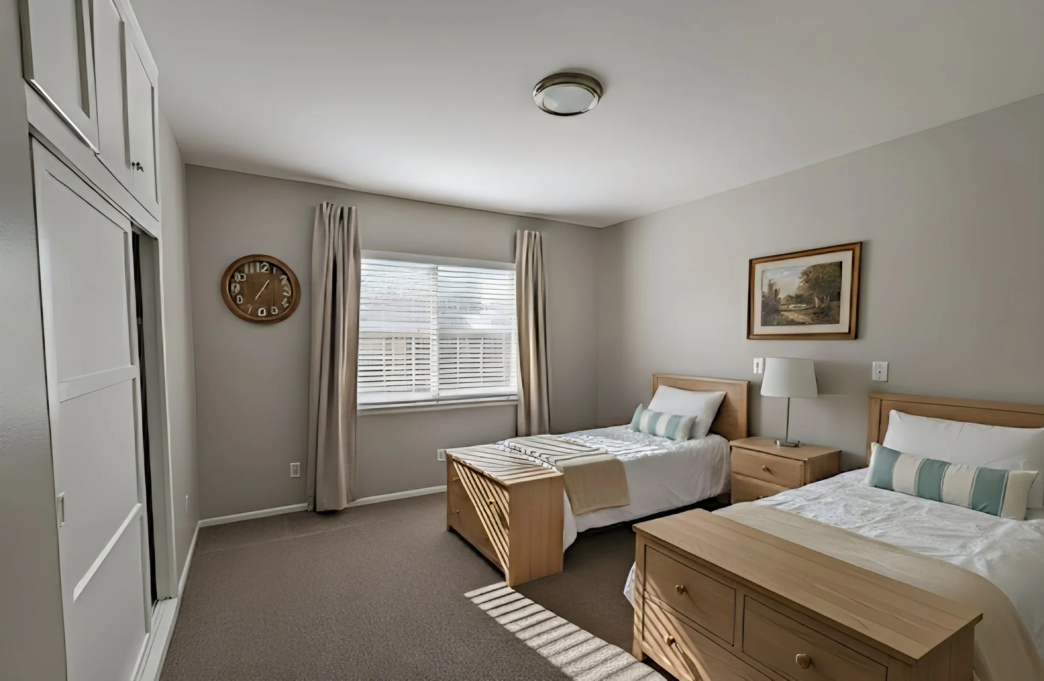 A bedroom with two twin beds, each with white linens and decorative pillows. A wooden nightstand with a lamp between the beds. A window with blinds and beige curtains, with sunlight casting shadows on the carpet. A framed landscape painting on the wall and a round wall clock.