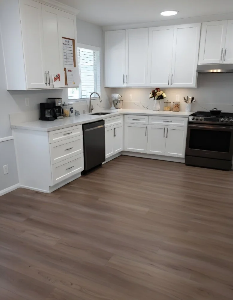 A modern kitchen with white cabinets, a stainless steel dishwasher, and a black oven. The countertop has coffee-making appliances, a bouquet of flowers, and kitchen utensils. There is a window with blinds and a small bulletin board on the wall.