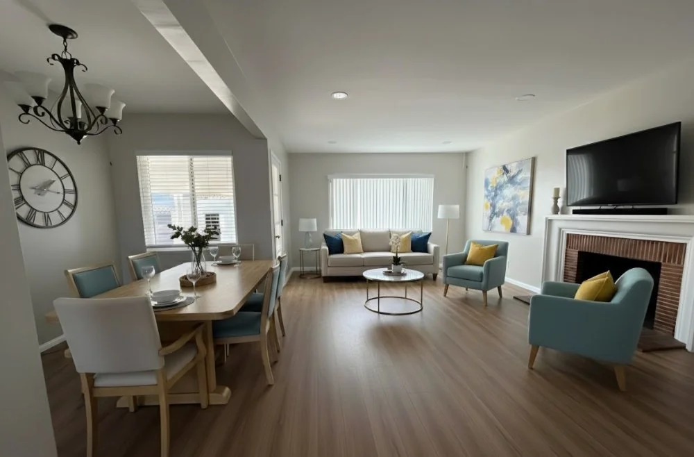 Living room with a dining area, featuring a wooden table with six chairs, a large wall clock, a sofa with multiple throw pillows, two blue armchairs with yellow pillows, a fireplace with a flat-screen TV above, and windows with blinds.