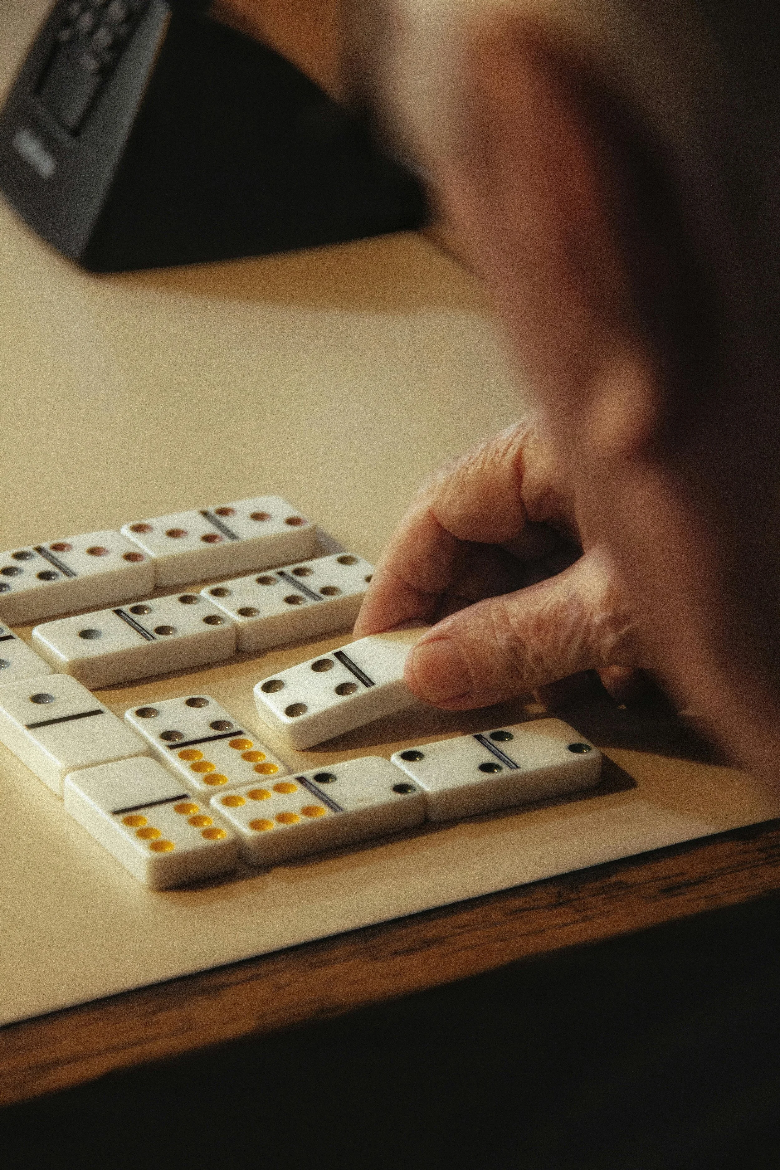 Close-up of a person playing a game of dominoes with white tiles featuring black and yellow dots on a wooden table.