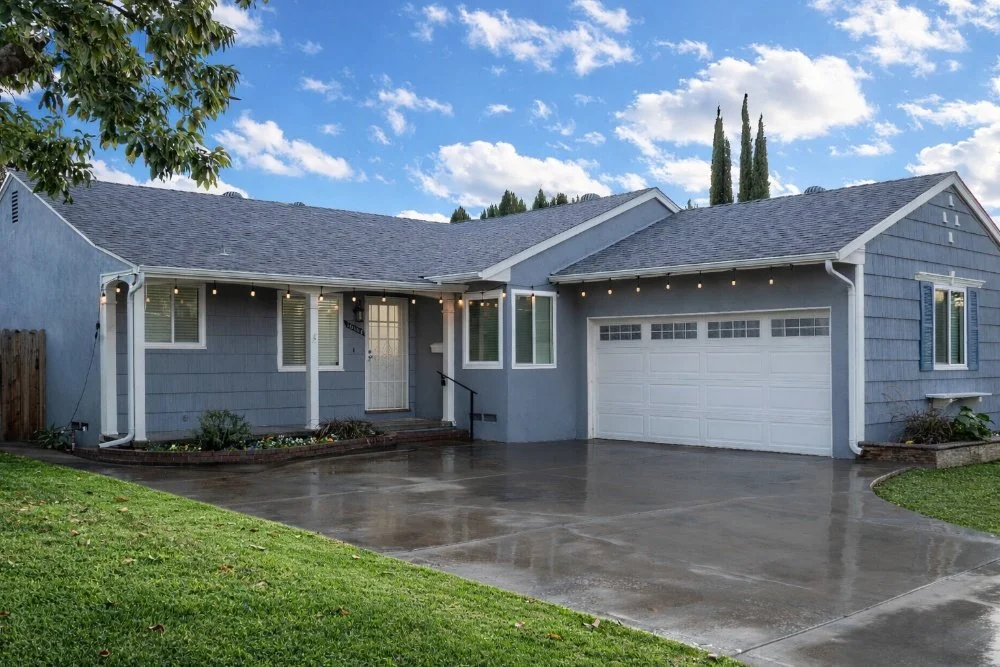 A blue single-story house with white trim, a front porch, attached garage, and string lights hanging in the yard, under a partly cloudy sky.