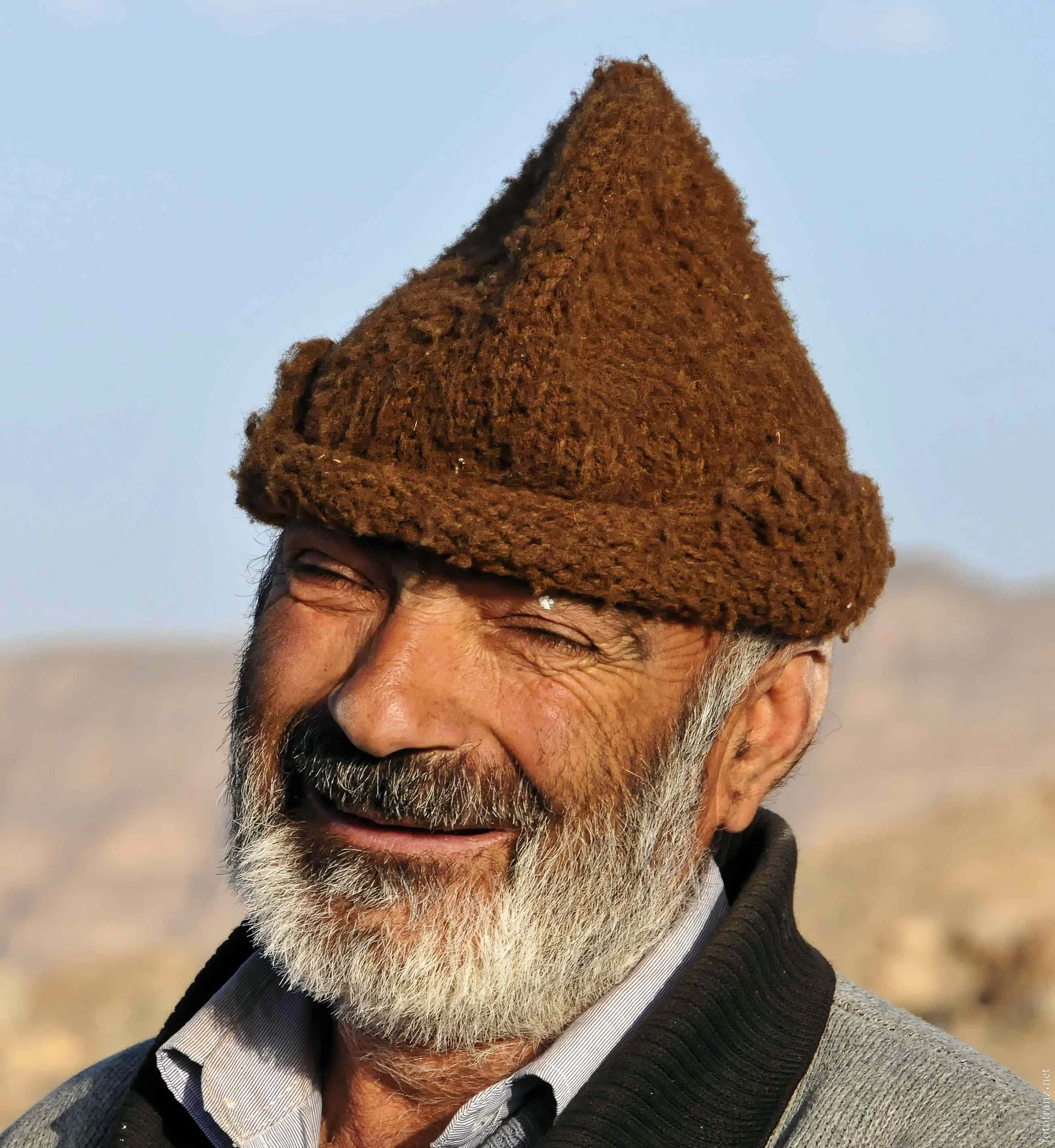 Ein lächelnder älterer Mann mit grauem Bart, trägt eine braune Wollmütze, schwarzes Oberteil und sieht fröhlich aus, im Hintergrund eine Berglandschaft bei sonnigem Wetter.