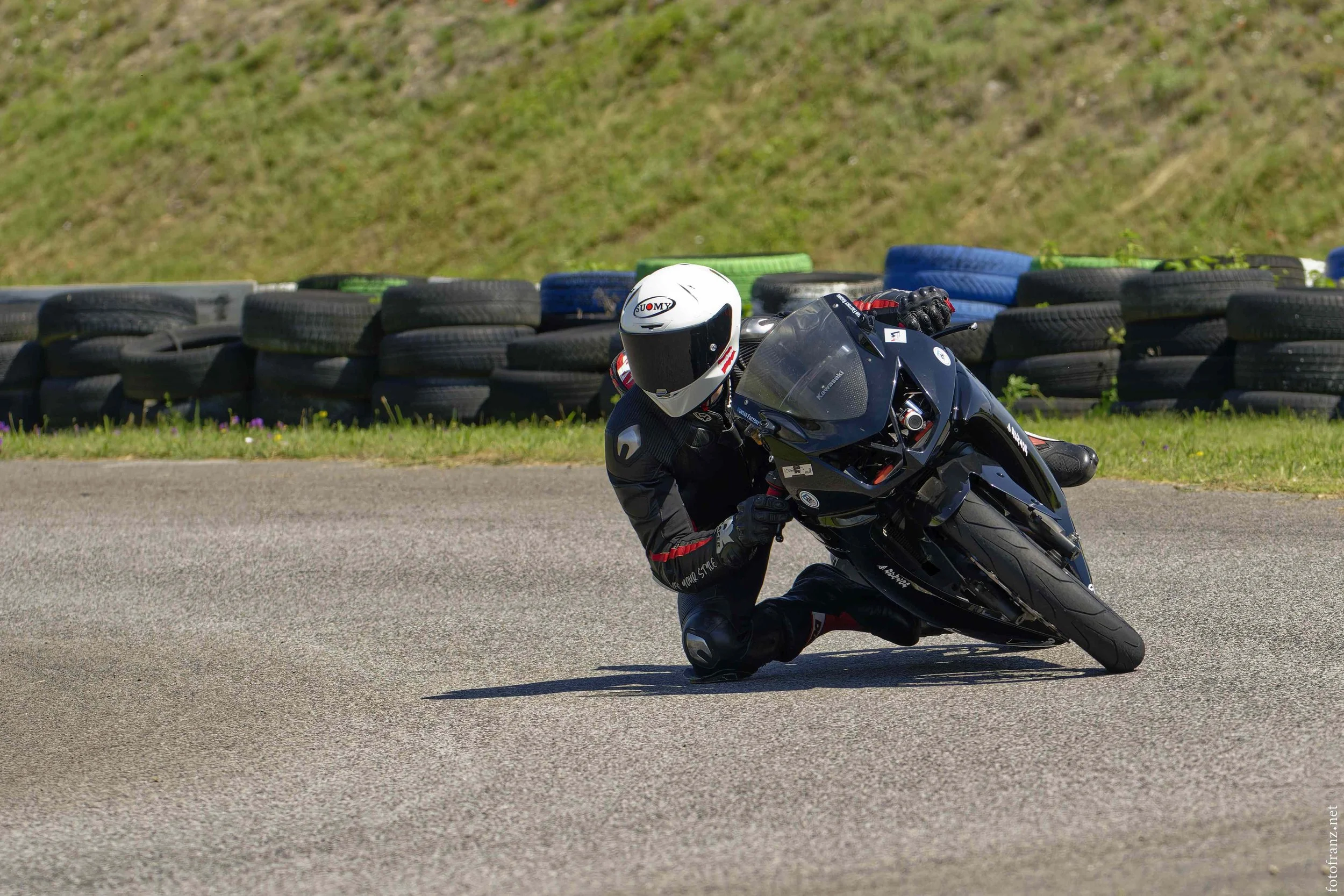 Motorradrennfahrer in schwarzer Kleidung und Helm auf einer überdachten Kurve auf einer Rennstrecke, umgeben von Reifenabsperrungen.