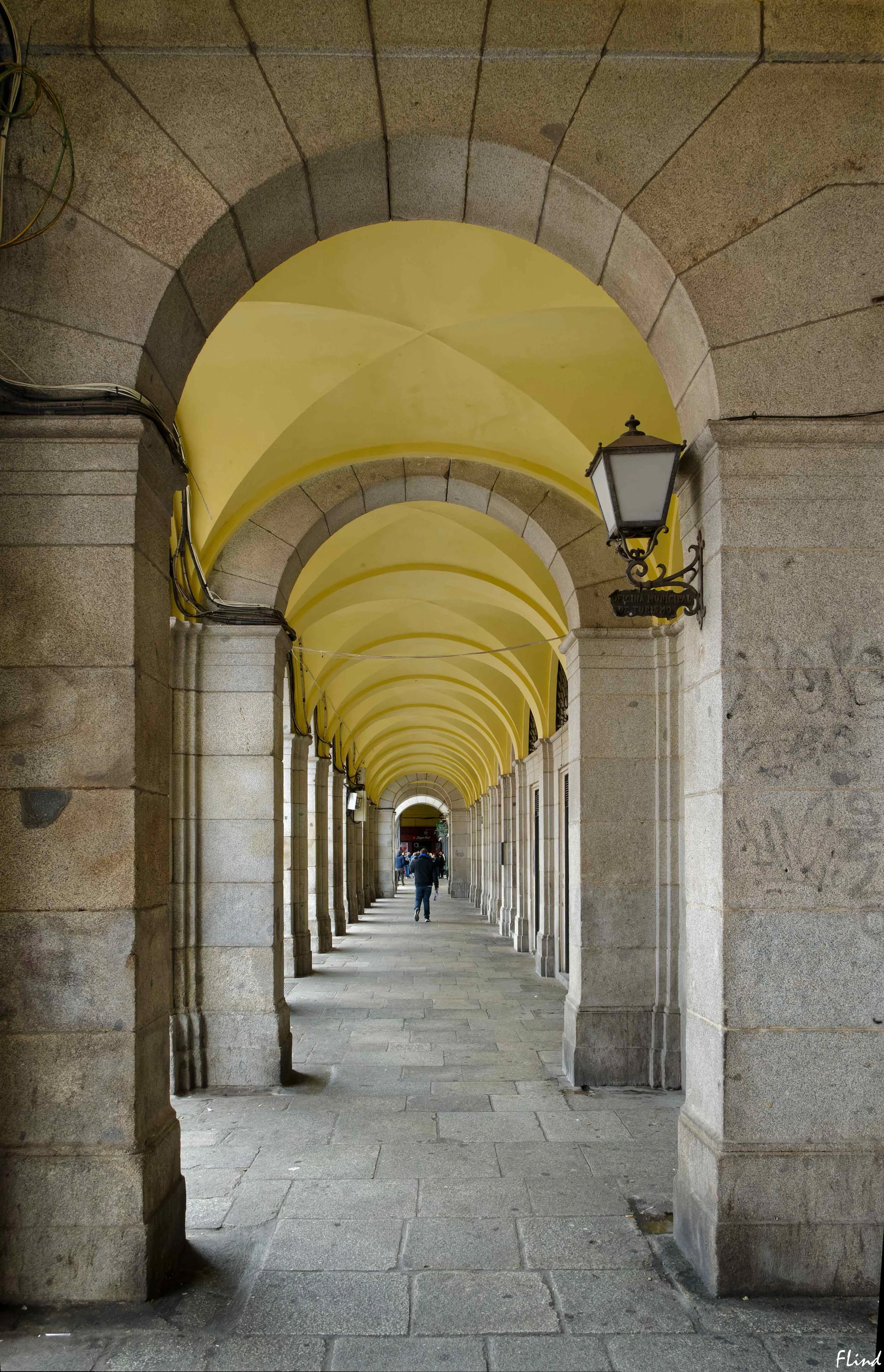Ein länglicher Fußgängertunnel mit hohen Steinbögen und hellgelber Decke, beleuchtet mit antiken Laternen, durch den Menschen gehen.