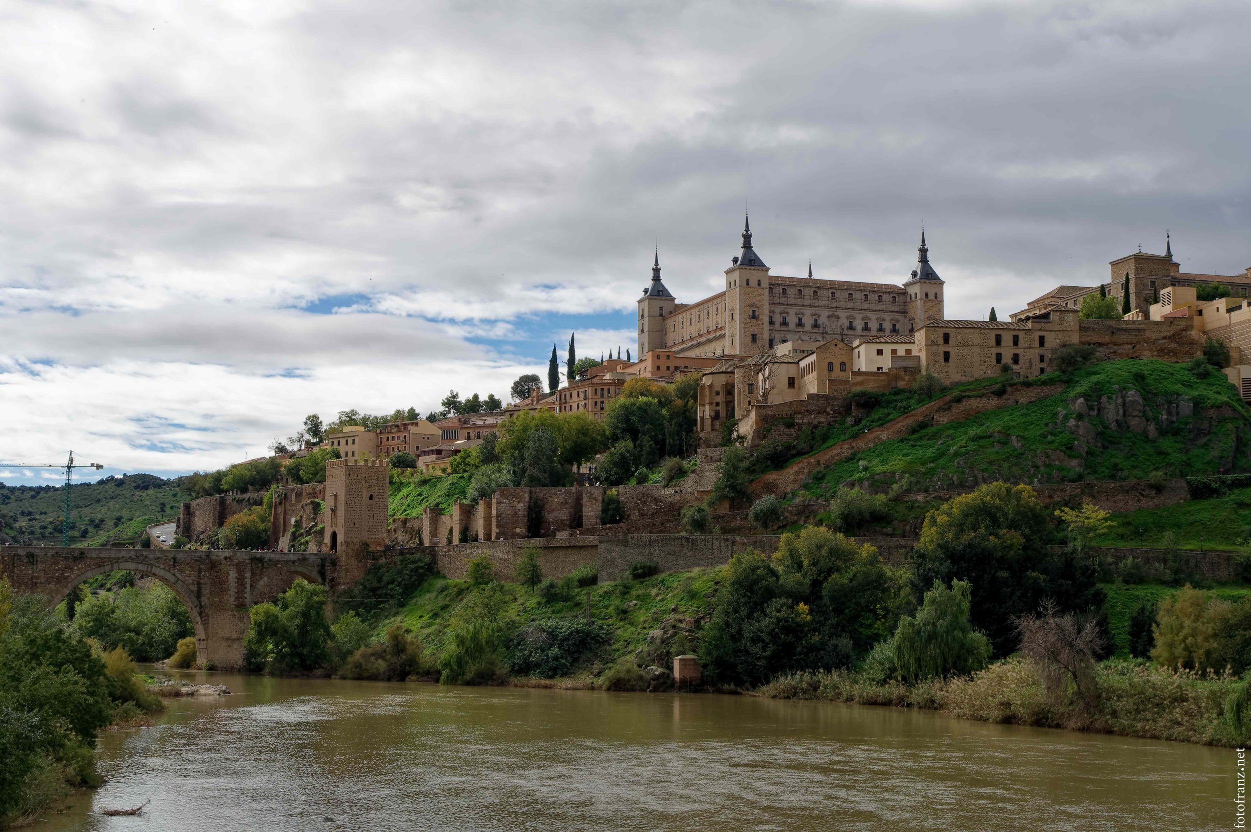 Historische Burg auf einem Hügel mit Fluss im Vordergrund und bewölktem Himmel.