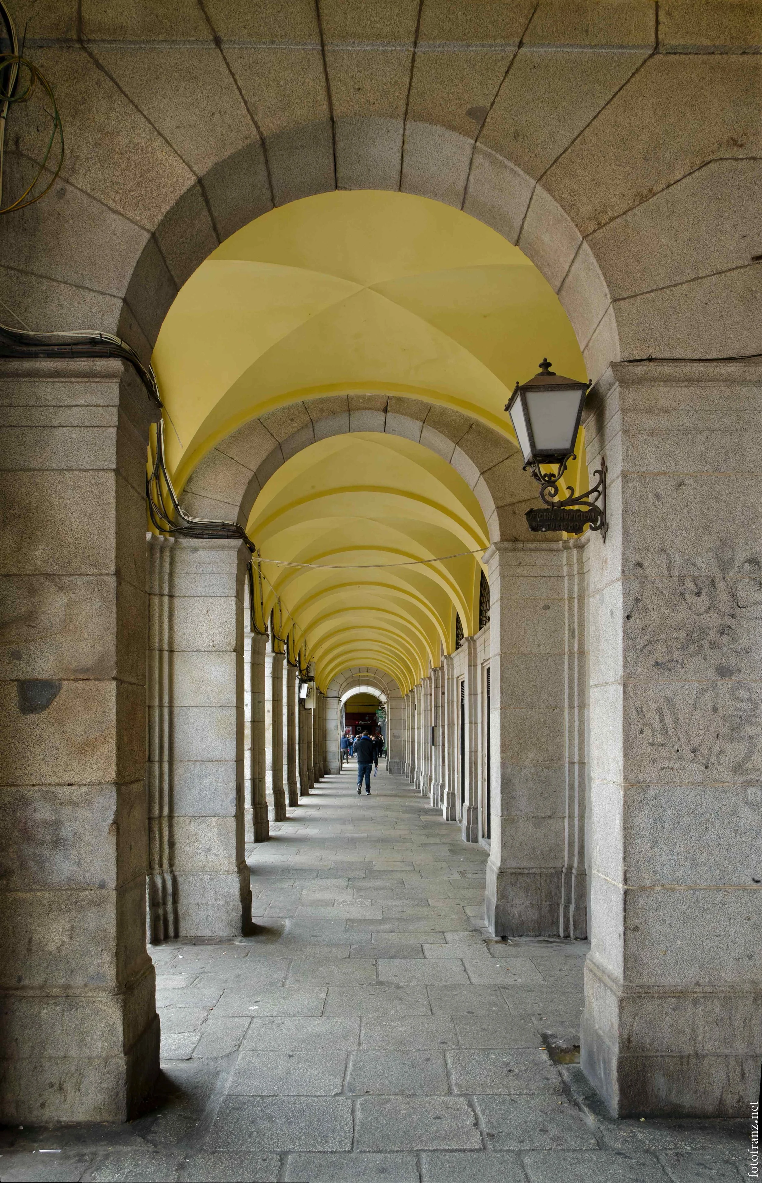 Ein langer Arkadengang mit gewölbter Decke und Laternen an den Säulen, Menschen laufen durch den Gang, im Hintergrund sichtbar.
