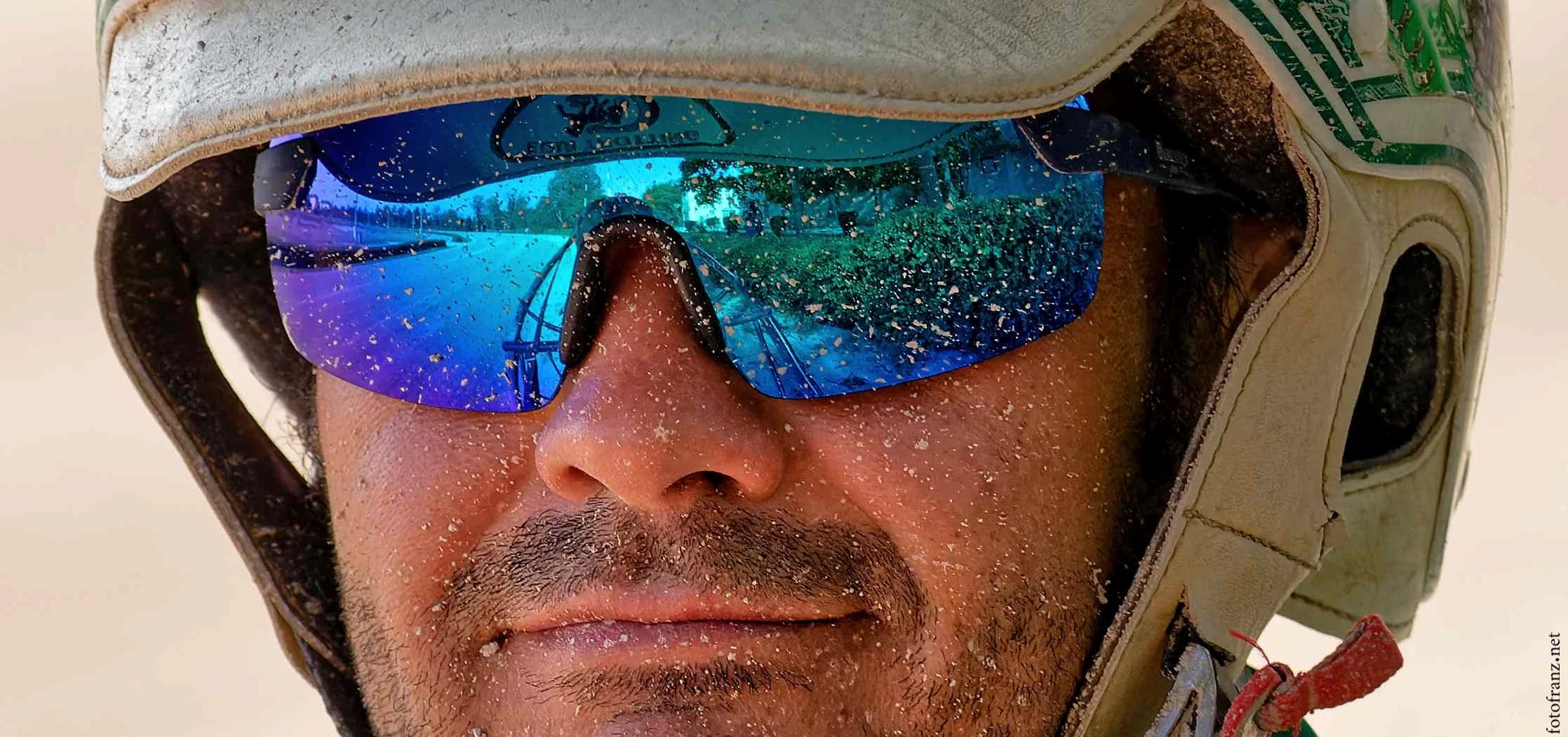 Nahaufnahme eines Mannes mit Sommerhelm, reflektierende Motorradbrille, Sand und Staub im Gesicht, im Hintergrund eine Wüstenlandschaft.