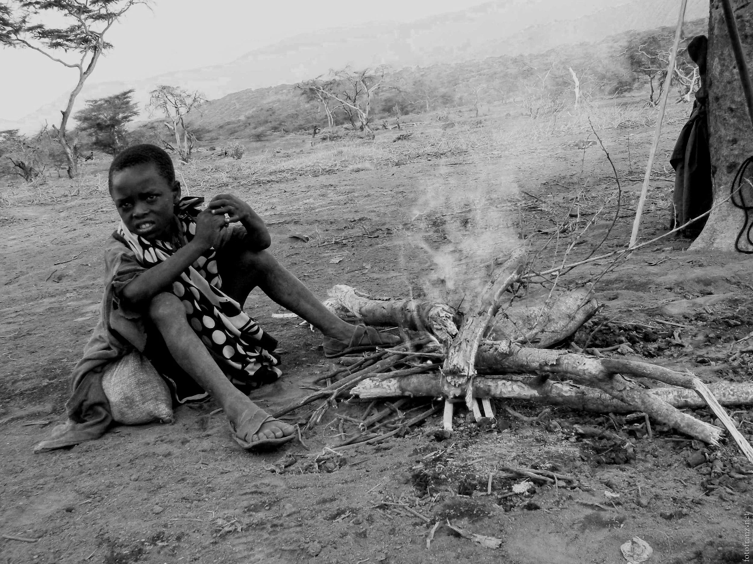 Kleiner Junge sitzt neben einem Feuer, das aus brennenden Ästen und Zweigen besteht, in einer trockenen, kargen Landschaft mit wenigen Bäumen im Hintergrund.