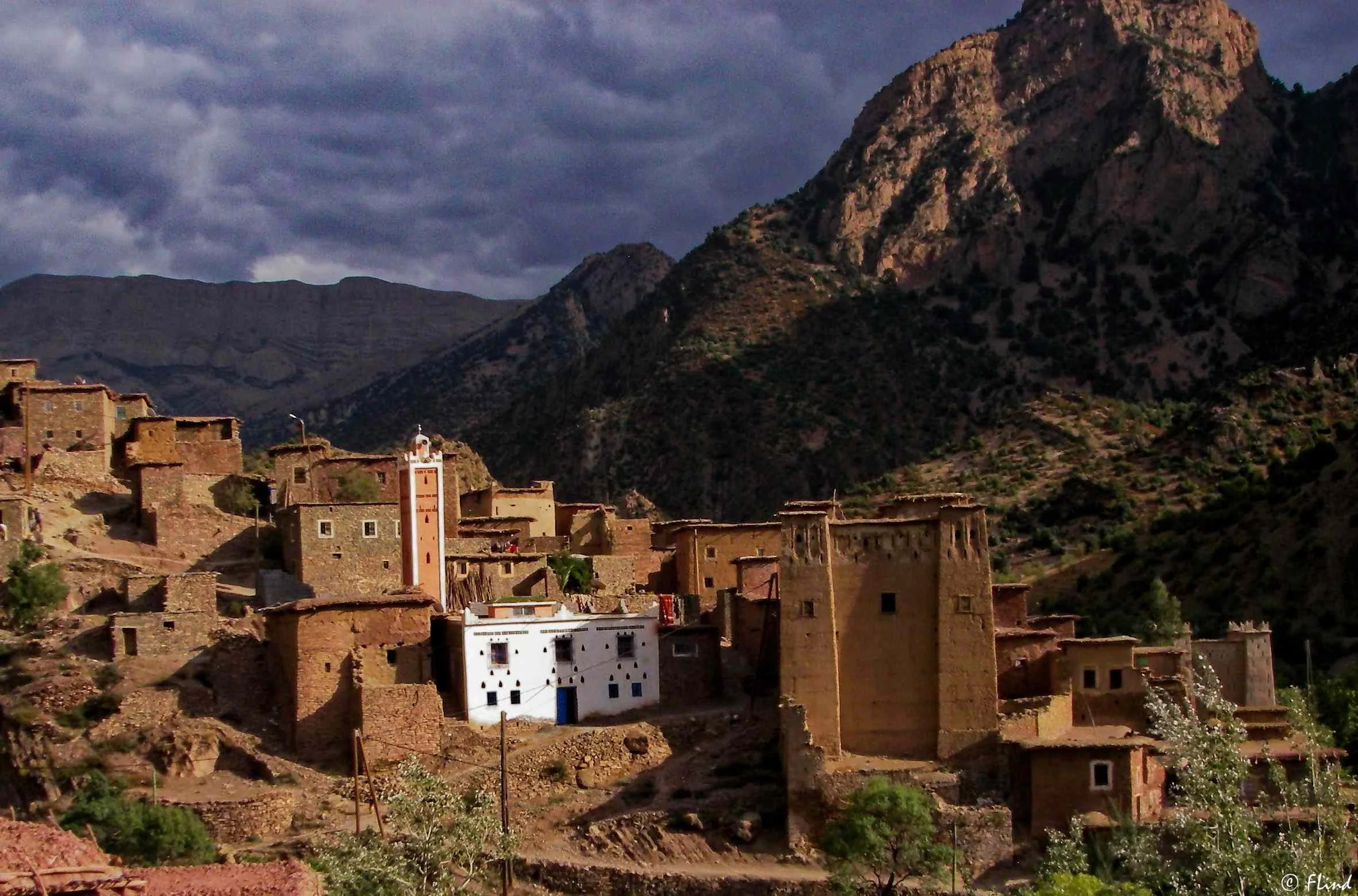 Bergdorf mit traditionellen Lehmbauten und einer Moschee im Berghang, umgeben von steiler Berglandschaft unter bewölktem Himmel in Marokko.