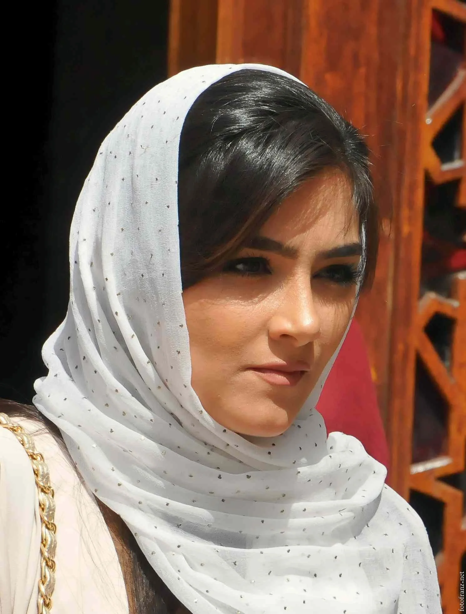 Nahaufnahme einer Frau mit schwarzem Haar, die einen weißen Kopftuch mit kleinen goldenen Punkten trägt, im Hintergrund Holzregal.