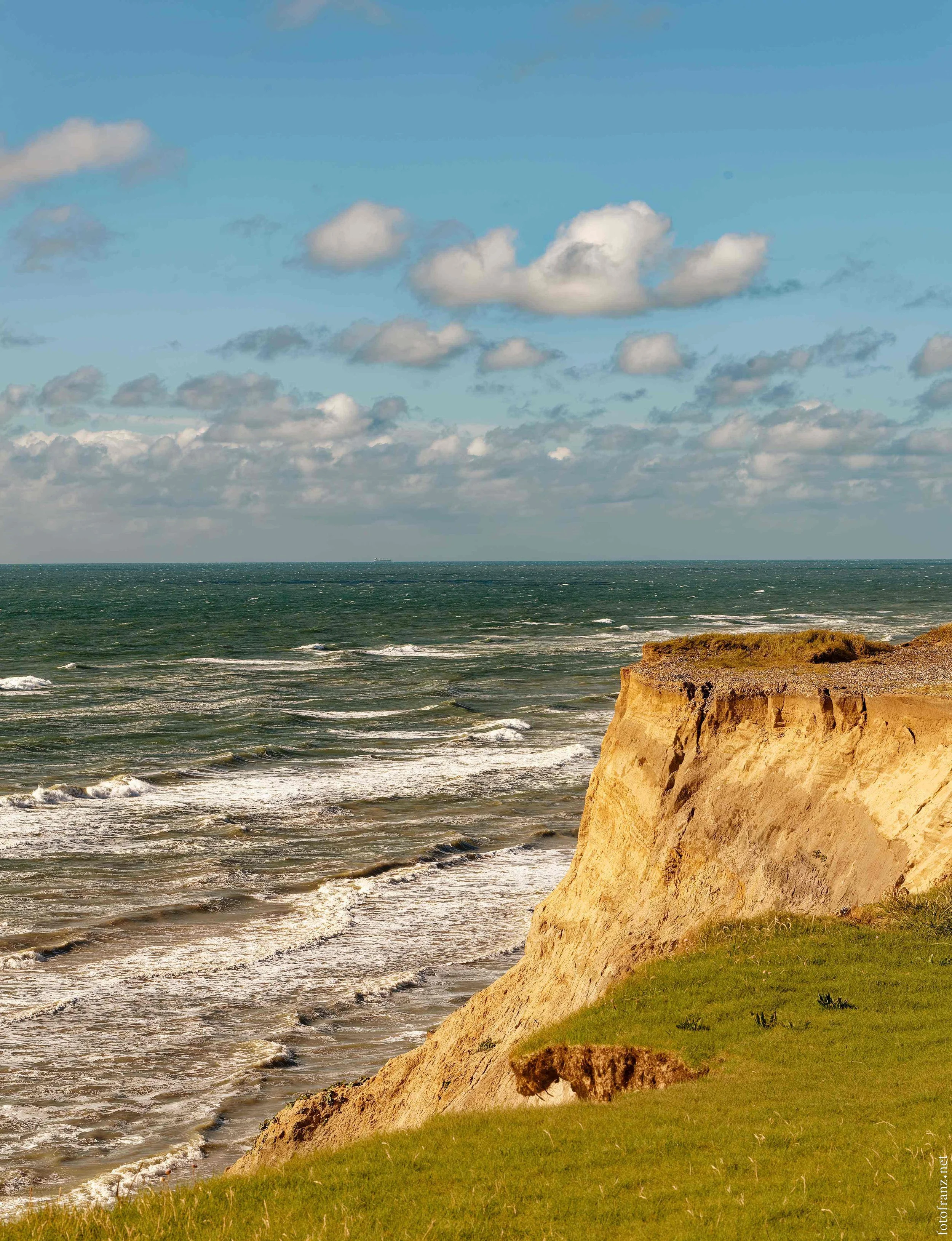 Klippen an einer steil abfallenden Küste mit Gras im Vordergrund, das Meer mit Wellen im Hintergrund und ein blauer Himmel mit Wolken.