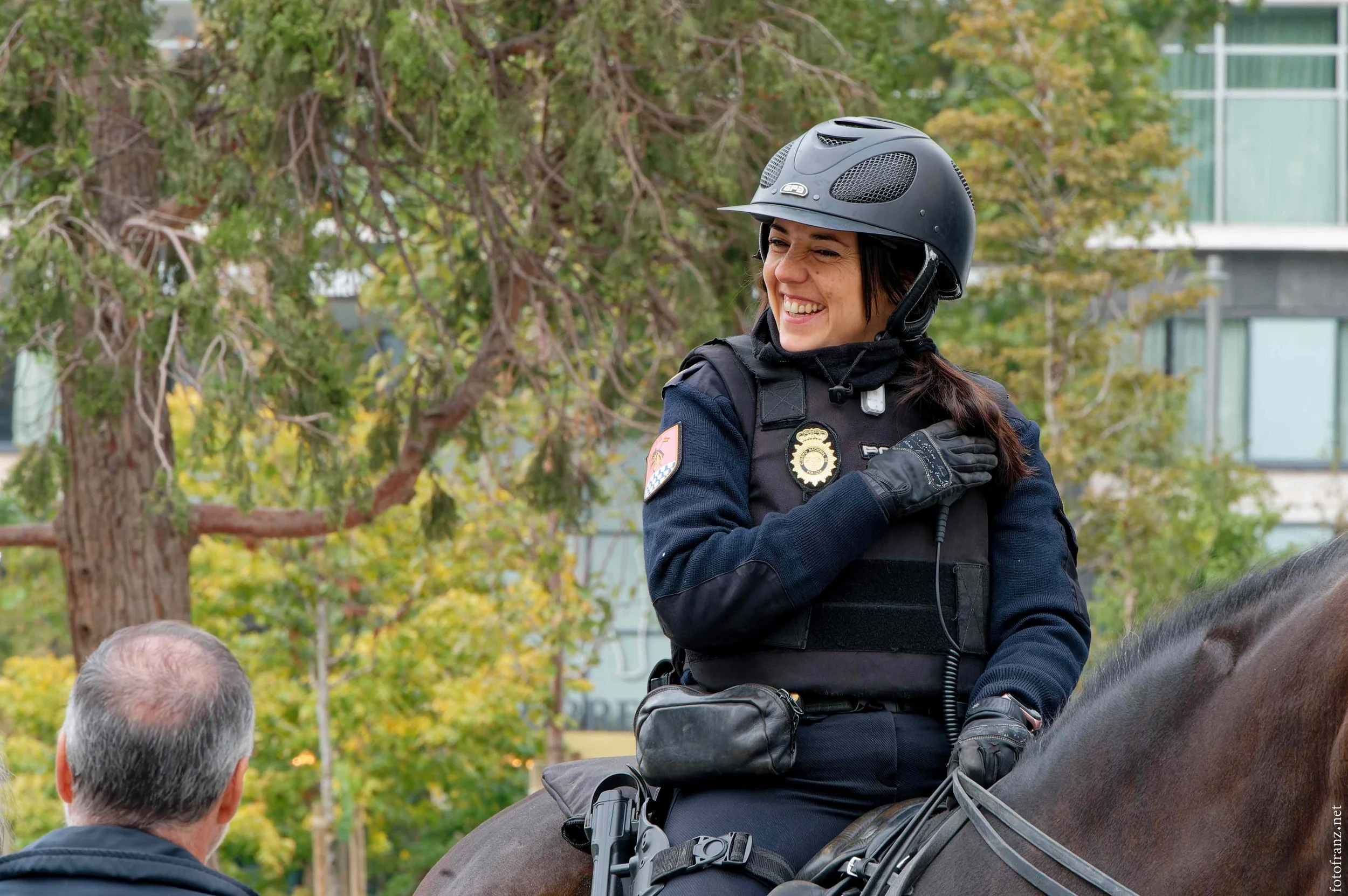 Eine lachende Polizistin trägt einen Helm und sitzt auf einem Pferd in einem Park, während sie mit einem Mann spricht.