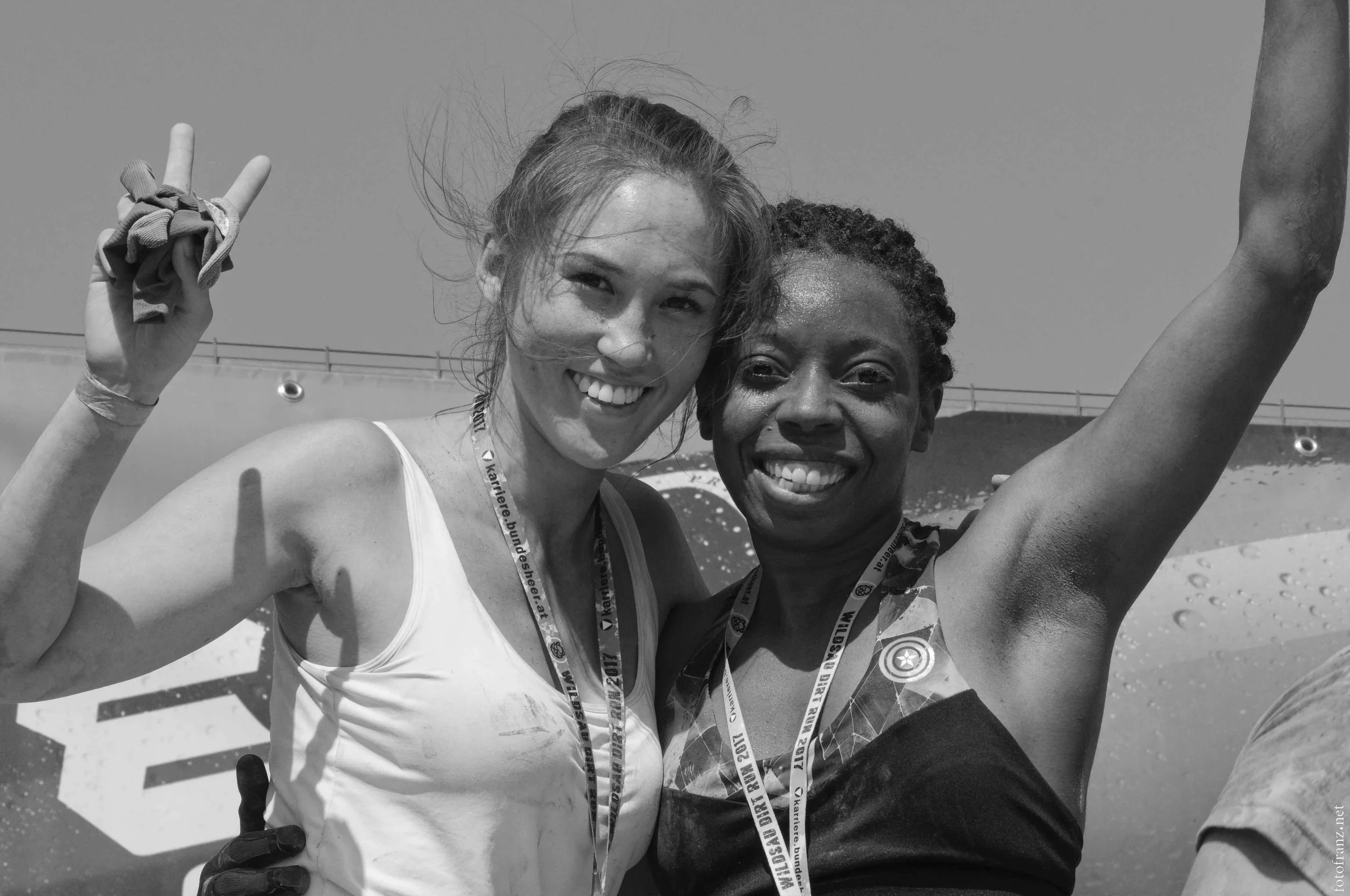 Zwei lächelnde Frauen beim Siegerfeier, eine mit weißem Sporttop, die andere mit dunkelblauem Top, beide tragen Medaillen, entspannter Moment nach einem Wettkampf im Freien.