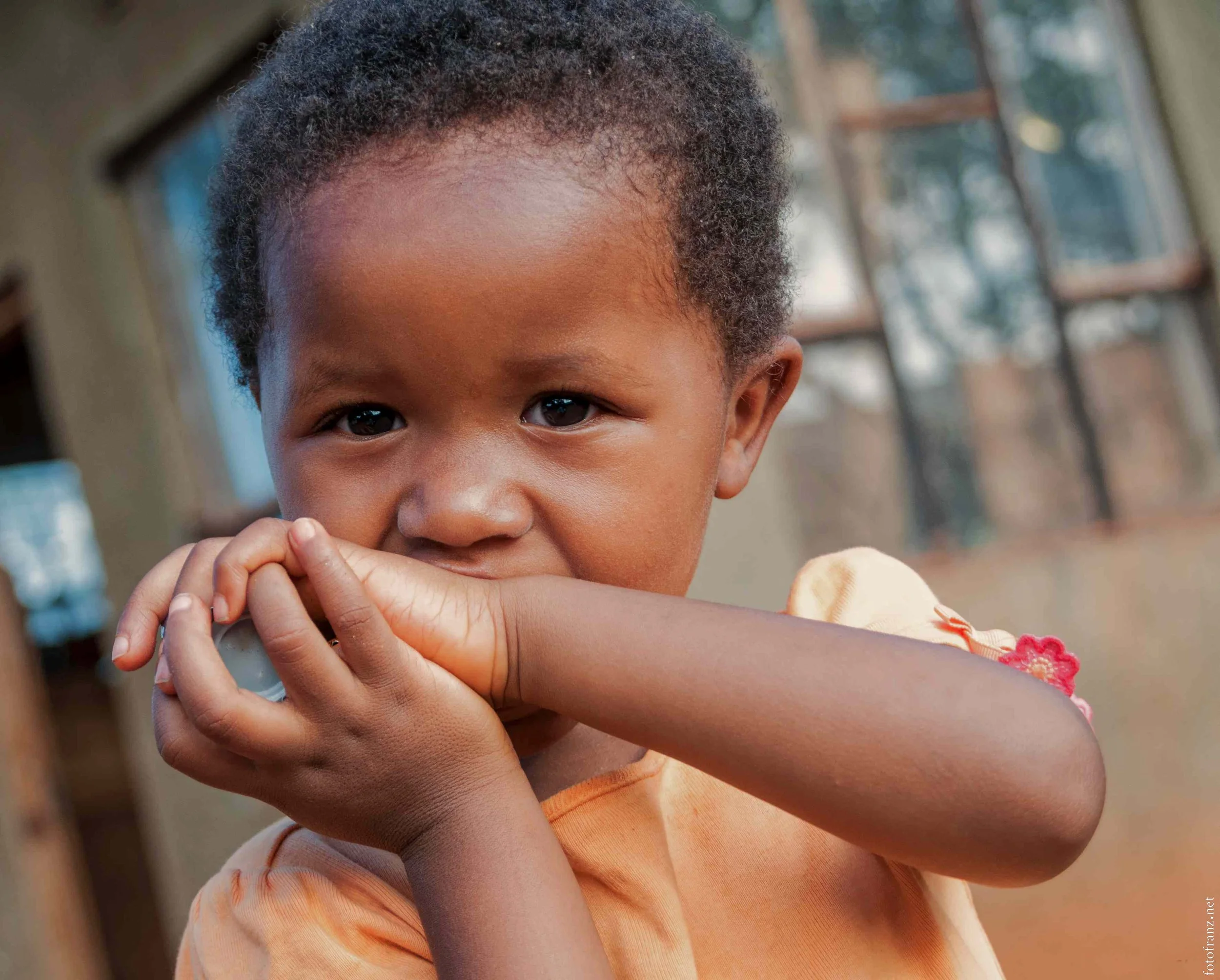 Ein junges Mädchen mit dunkler Haut und curly Haaren, das im Freien nahe einem Haus mit großen Fenstern steht, während sie in die Kamera schaut und die Arme vor das Gesicht hält.