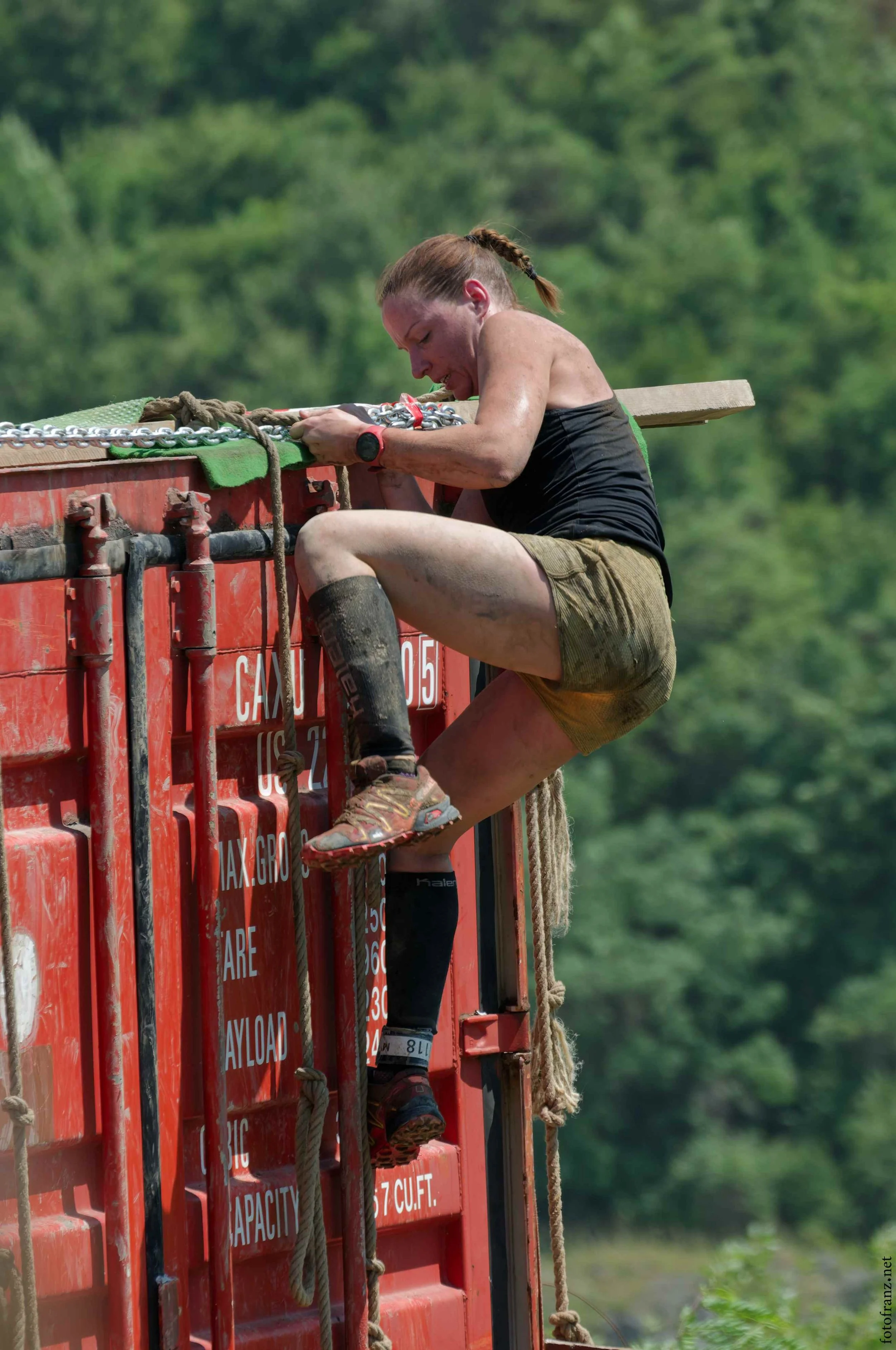 Eine Frau bei einem Hindernislauf, die auf einem roten Container hochklettert und sich festhält.