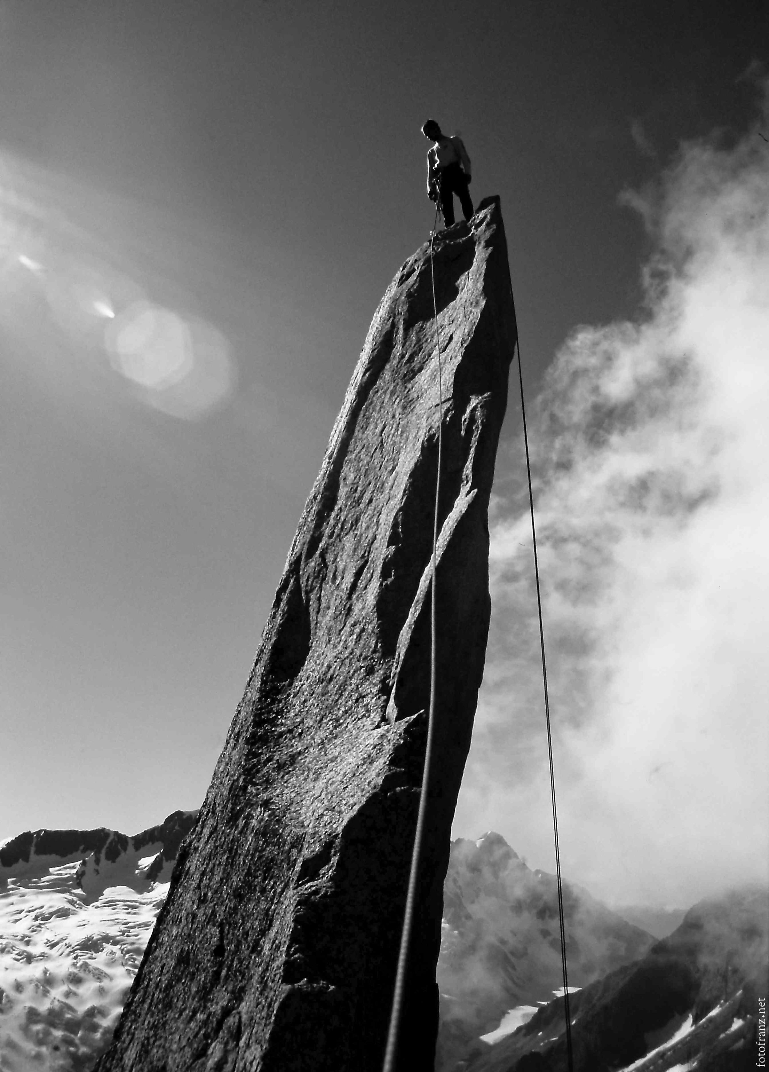 Kletterer steht oben auf einem schmalen, hohen Felsen mit einer Seilsicherung in den Bergen, im Hintergrund sind Schneegipfel und Wolken.