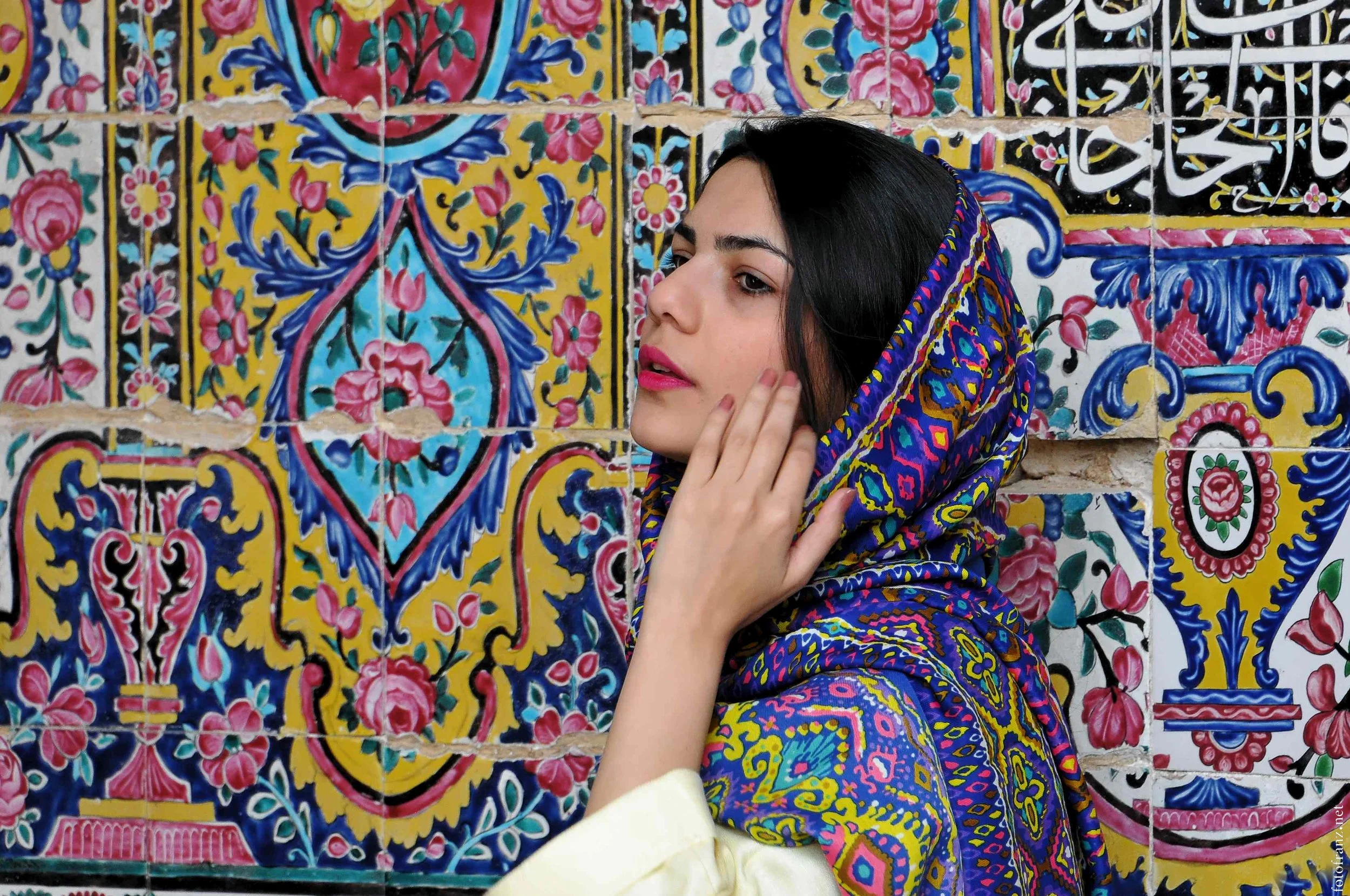 Junge Frau mit einem bunten schal, der über ihren Kopf drapiert ist, steht vor einer Wand aus kunstvoll verzierten, bunten Kacheln mit floralem und geometrischem Muster.
