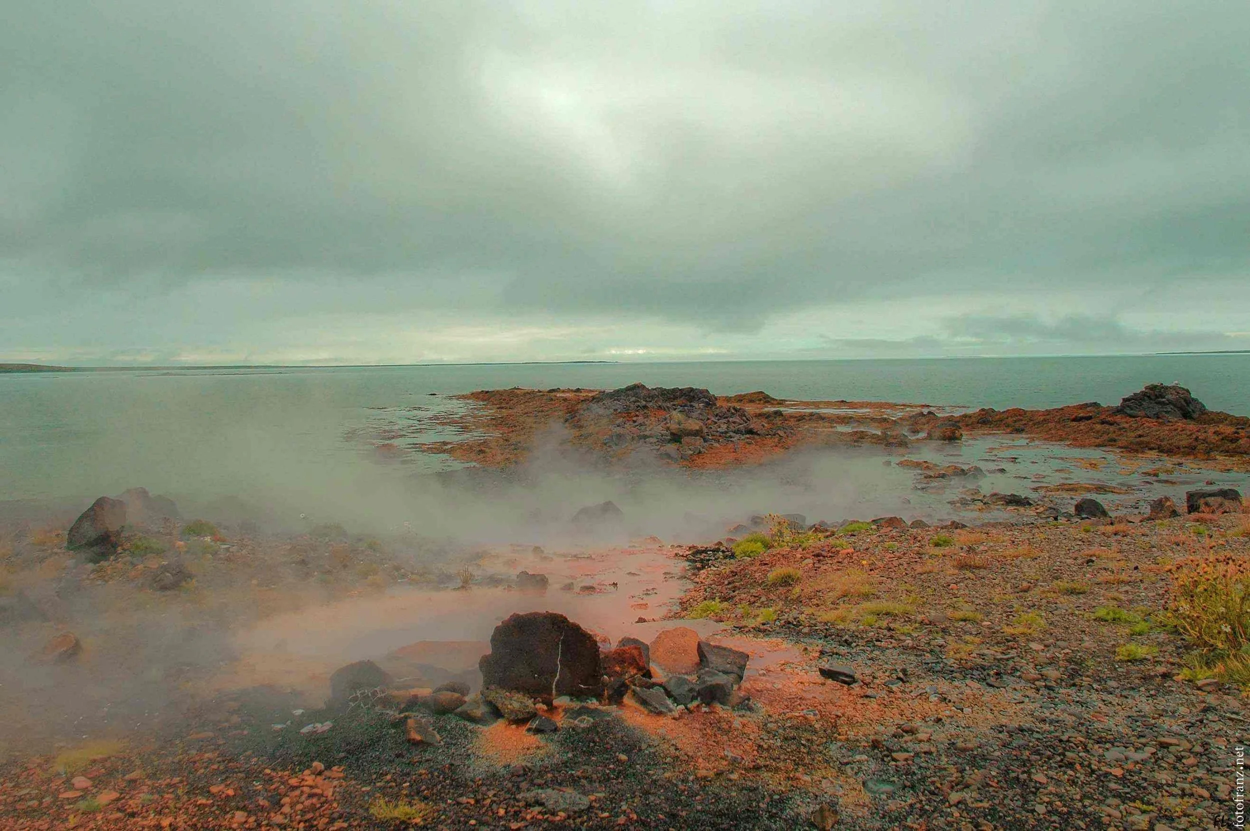 Vulkanischer Küstenabschnitt mit kochendem Wasser, dunklen Steinen und rauchenden mineralischen Quellen unter bewölktem Himmel.