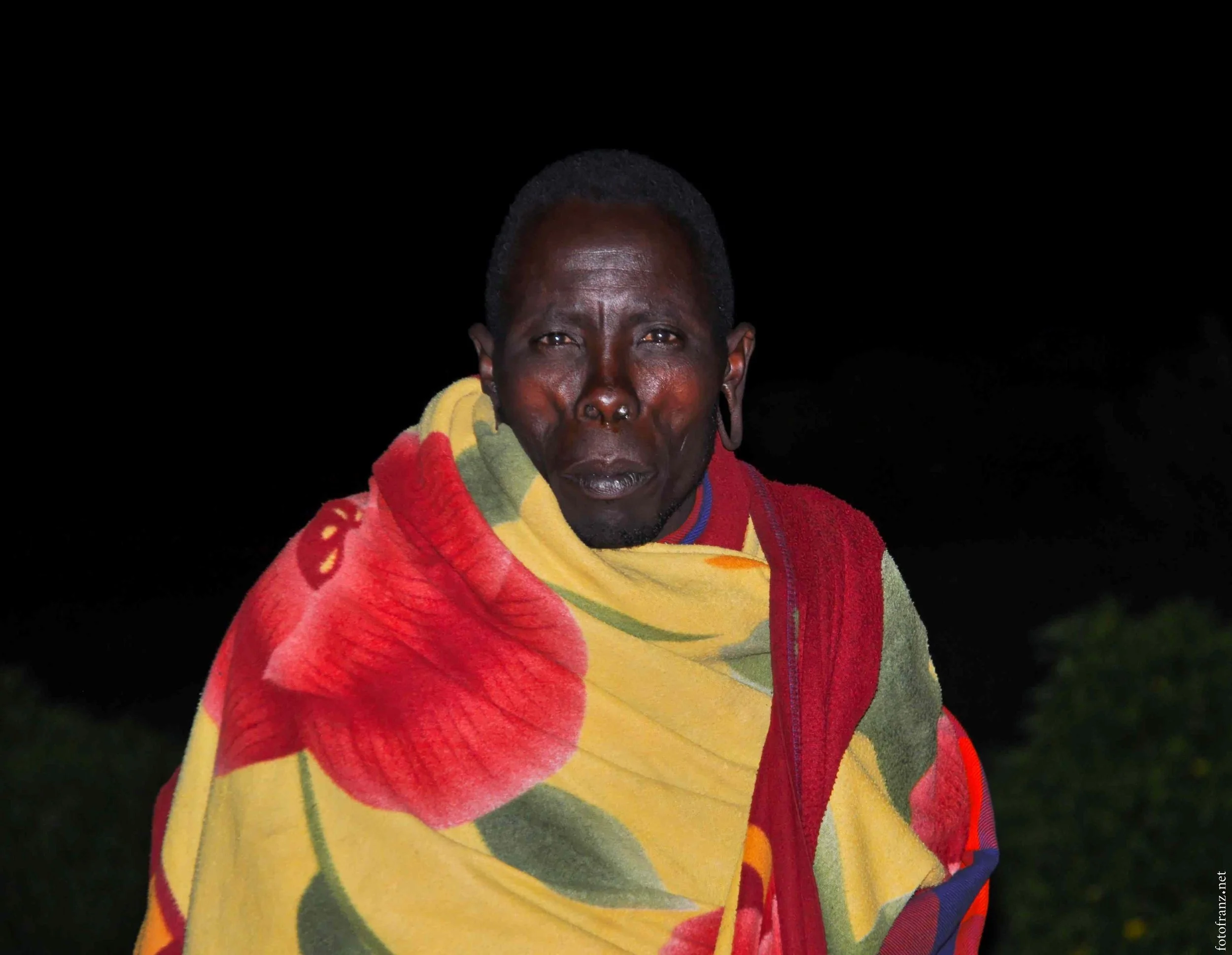 Eine Person mit dunkler Haut trägt einen bunten, floral gemusterten Mantel, der Gelb, Rot und Grün enthält. Die Person steht vor einem dunklen Hintergrund.