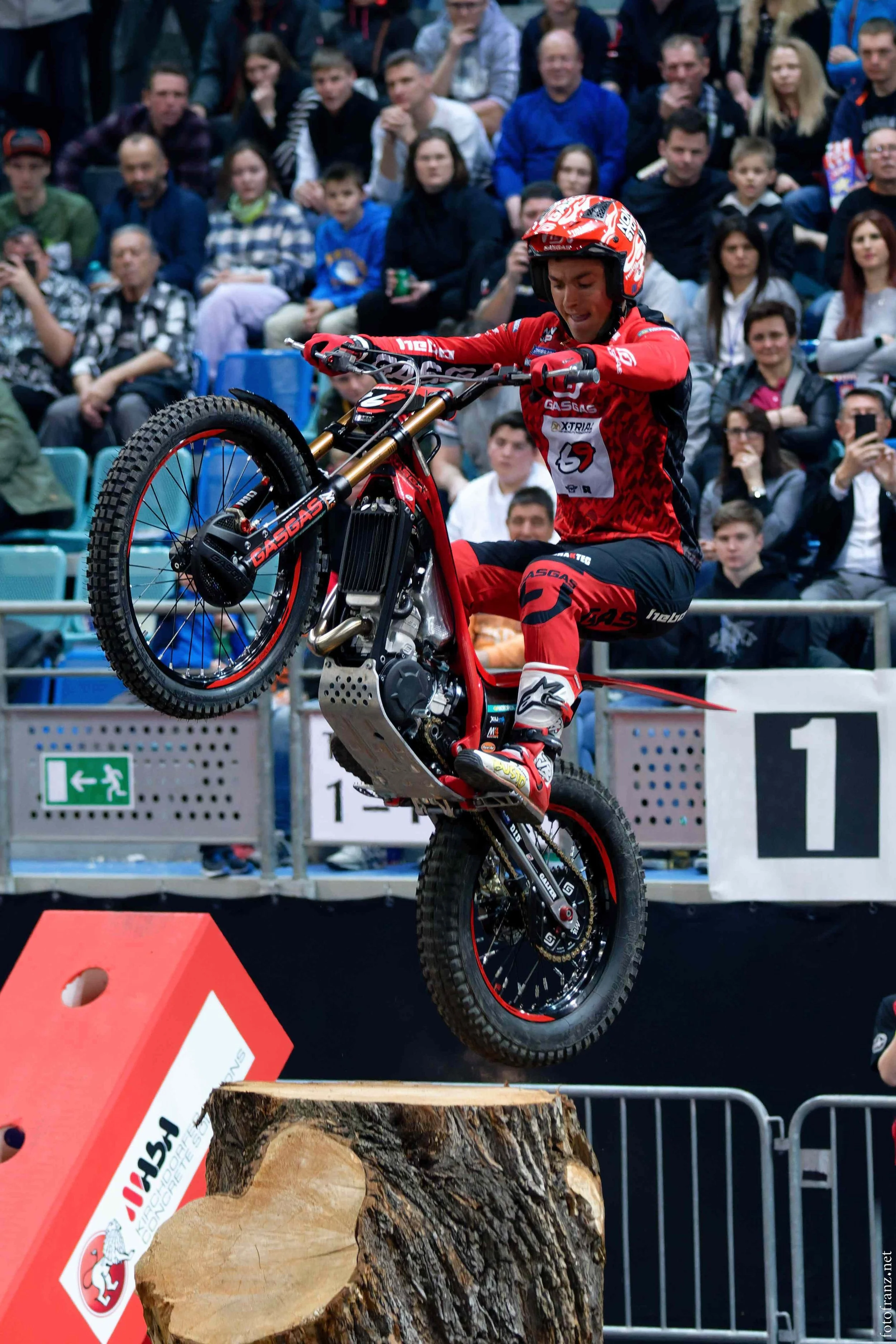 Motorradfahrer macht einen Wheelie während eines Wettbewerbs, im Hintergrund jubelnde Zuschauer.