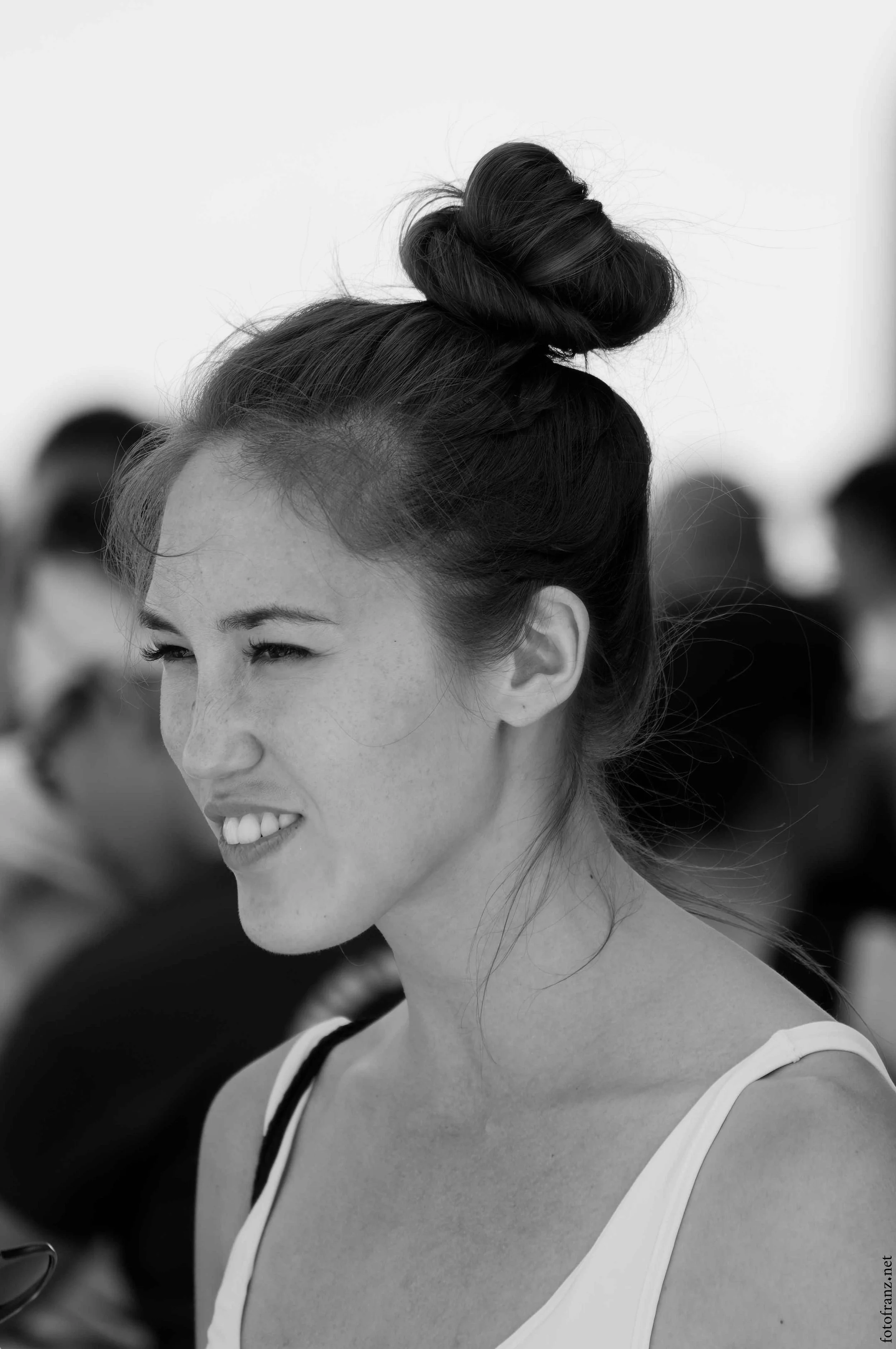 Schwarz-weiß-Foto eines lachenden Mädchens mit einem hohen Dutt und einem weißen Top, im Hintergrund sind andere Menschen sichtbar.