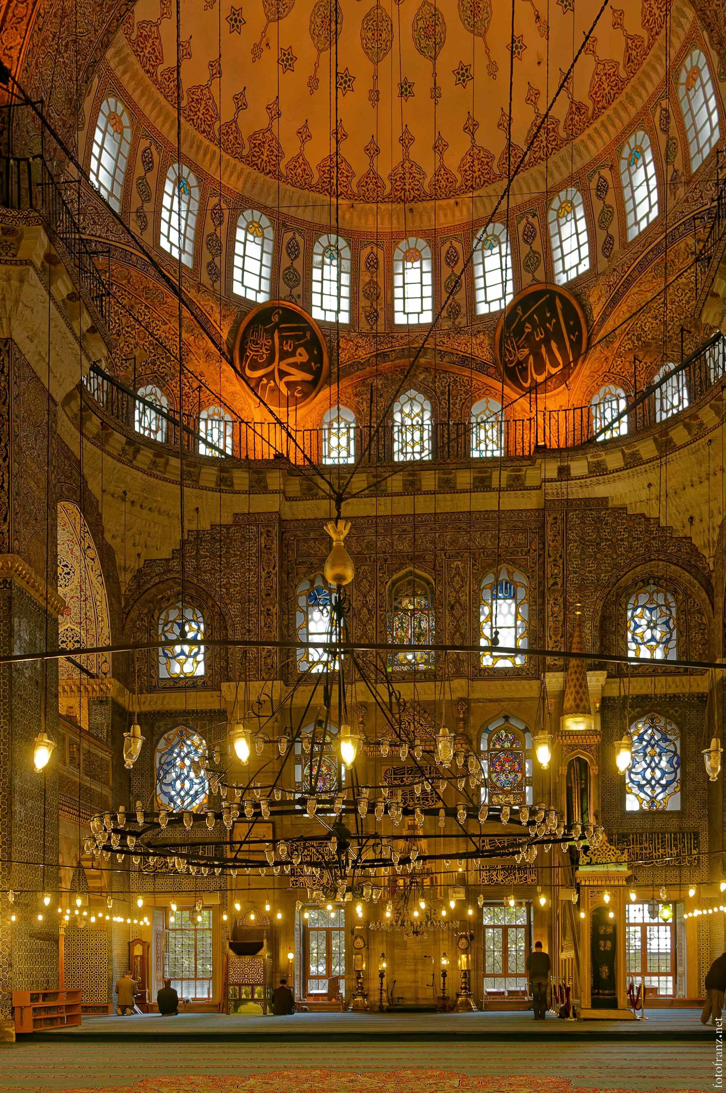 Innenaufnahme einer Moschee mit hohen Fenstern, Kalligraphie und kunstvollen Dekorationen, einige Personen beim Gebet.