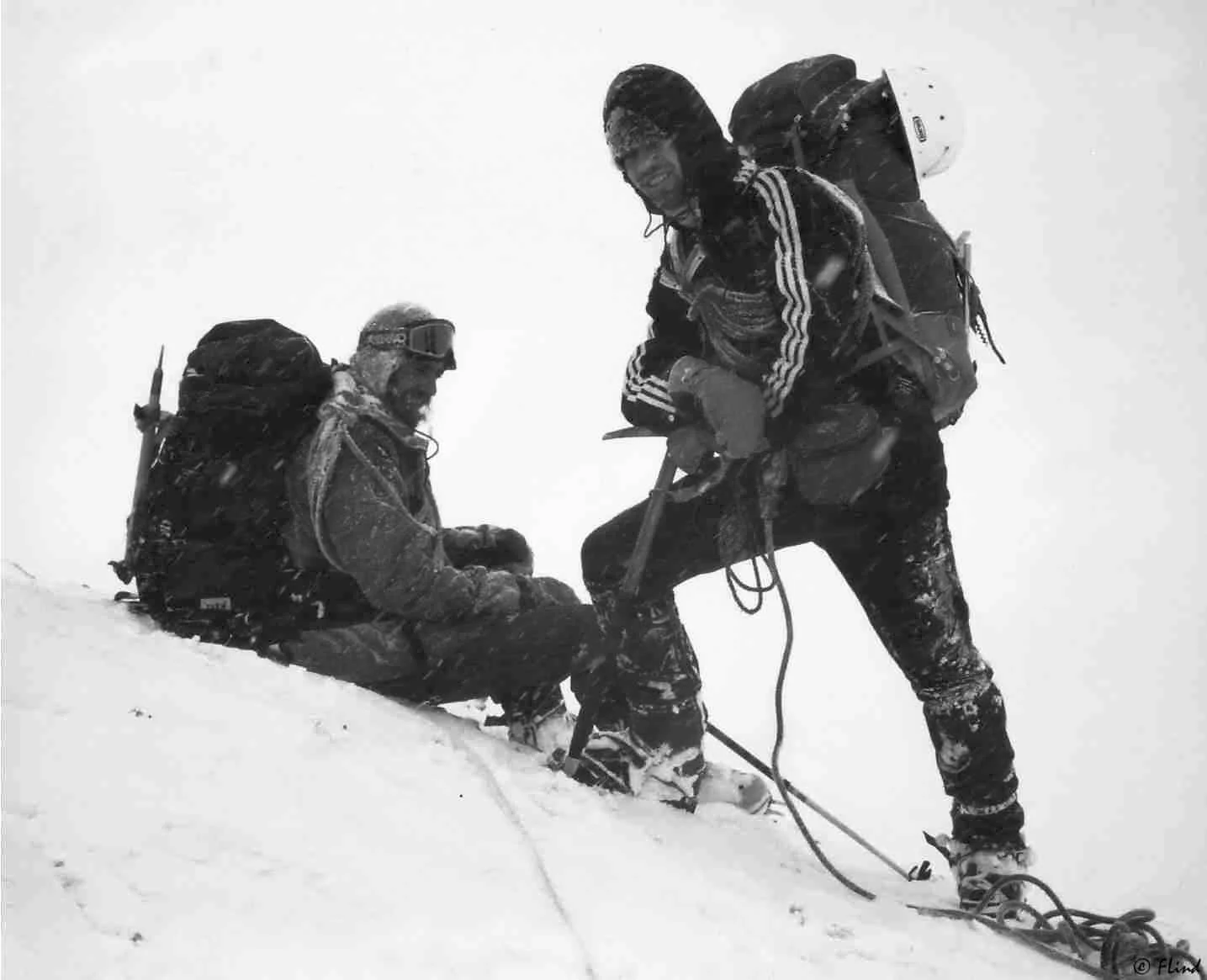 Zwei Bergsteiger im Schnee, einer sitzt, der andere steht mit Steigeisen und Eispickel, in starkem Schneefall.