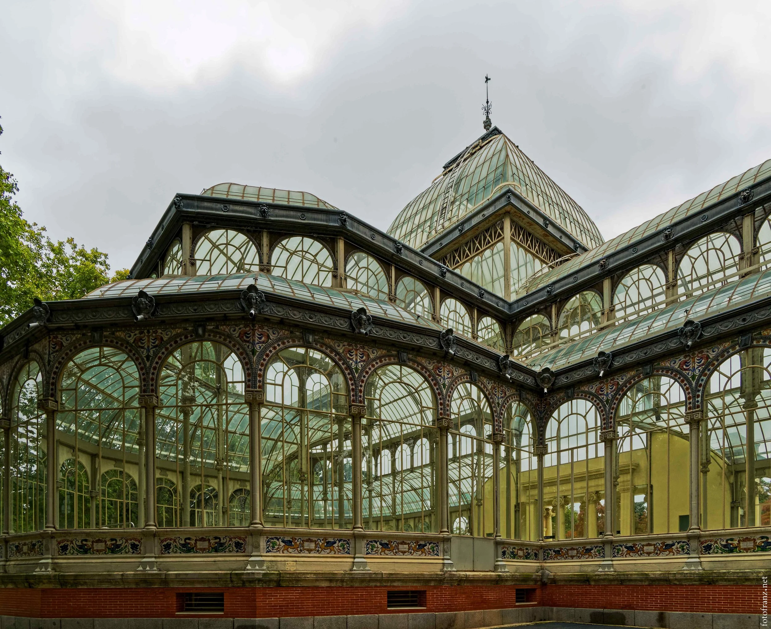 Ein prächtiges historisches Gewächshaus mit Glas- und Metallrahmen, verziert mit bunten Mosaiken, vor einem bewölkten Himmel.