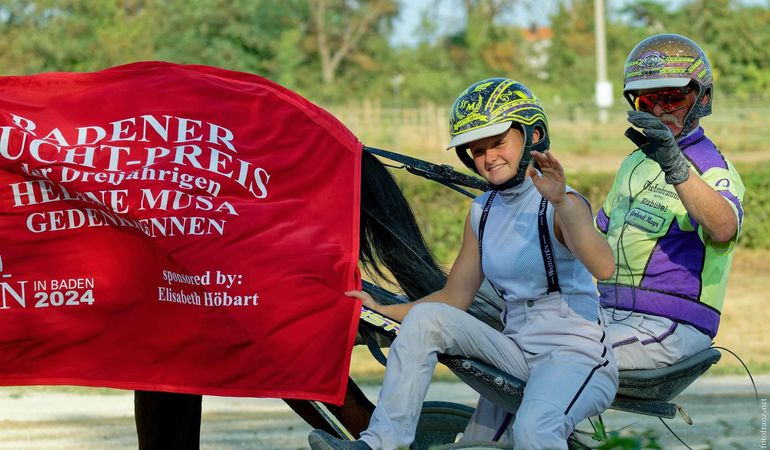 Ein junger Mädchen auf einem Pferd mit einem Erwachsenen im Reithelm, beide tragen Reiterkleidung, auf einem Feld mit Bäumen im Hintergrund, es ist sonnig.