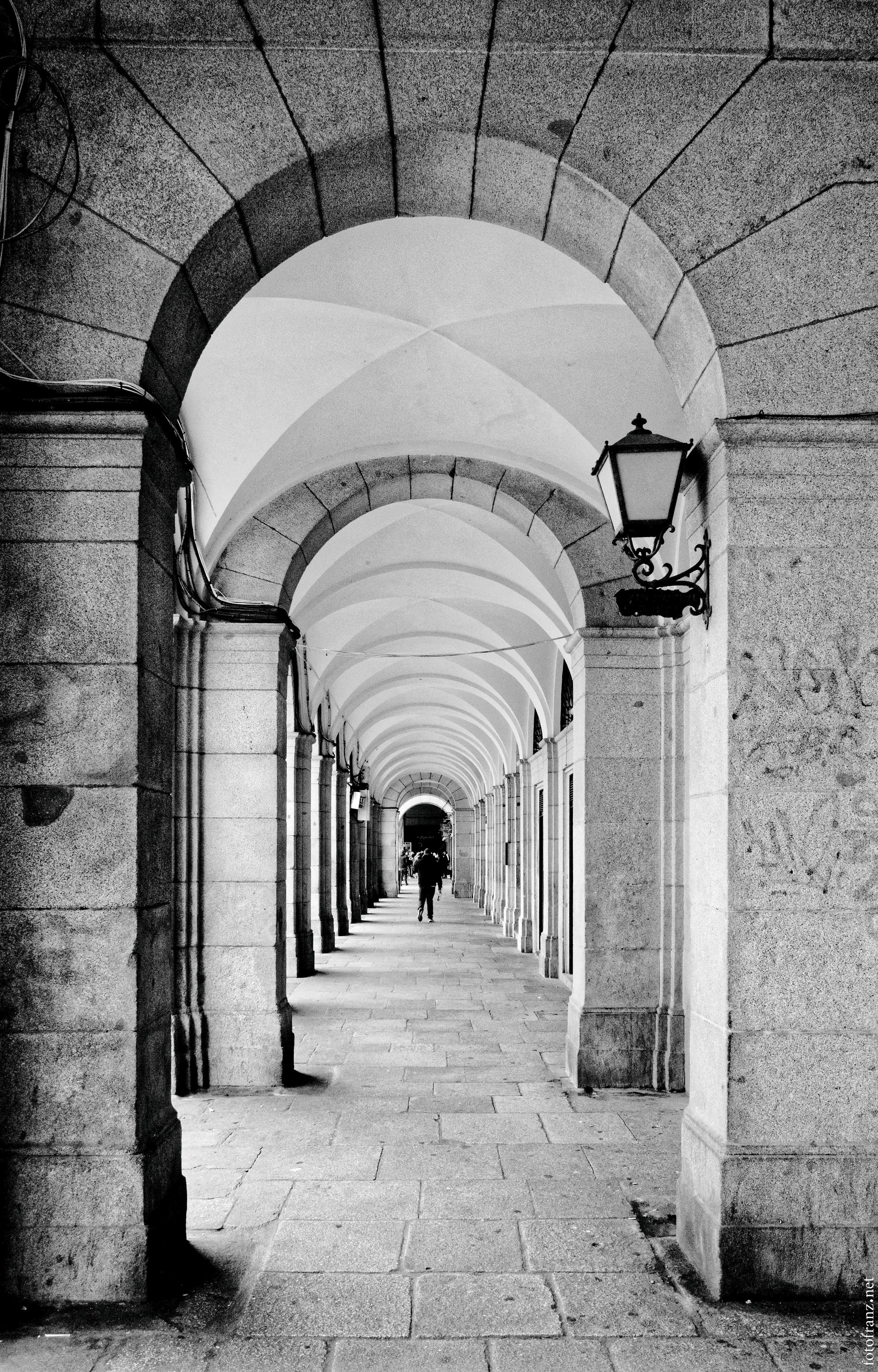 Blick durch eine historische Bogengang mit Laternen an den Wänden, im Hintergrund sind Menschen zu sehen.