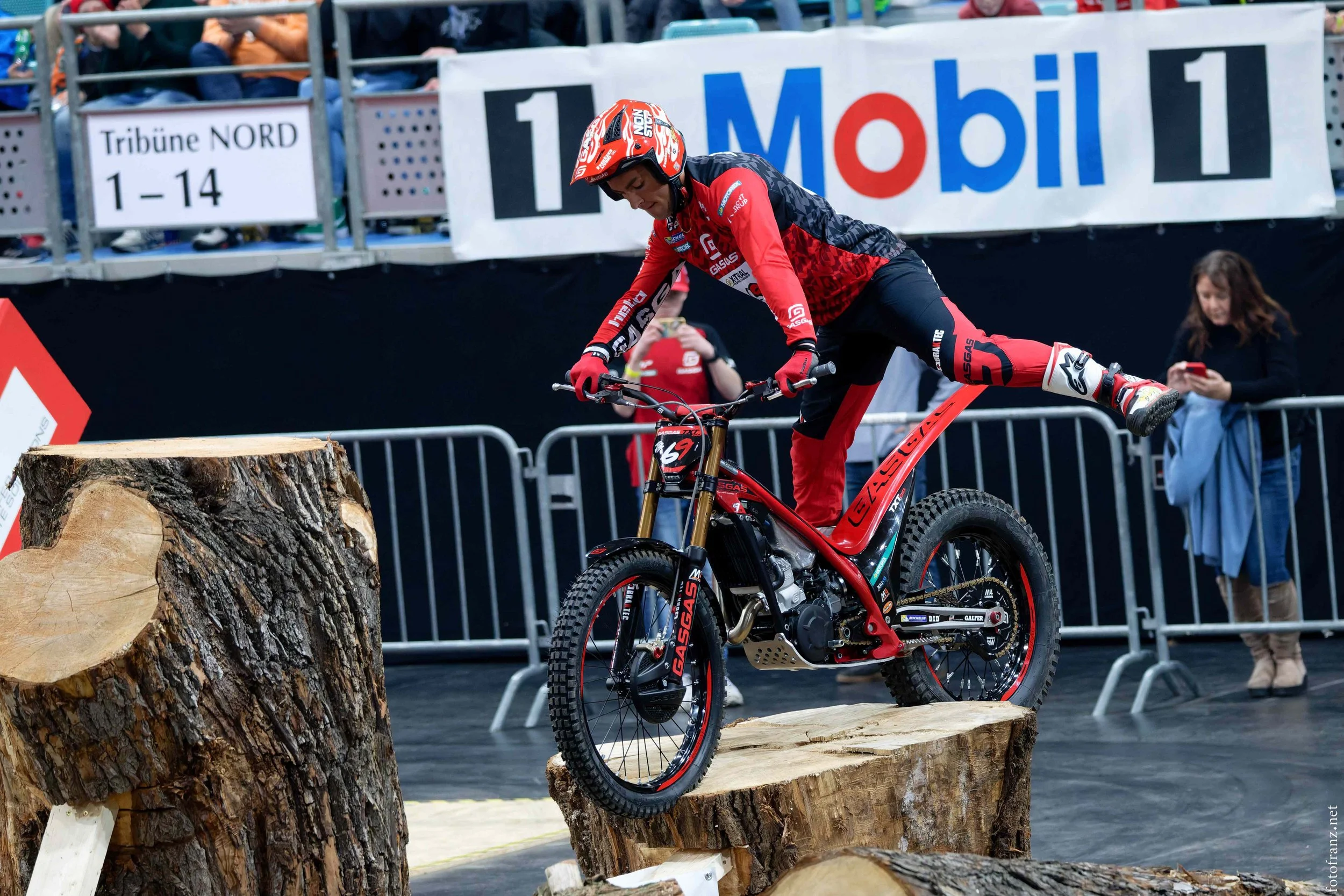 Ein Mountainbike-Fahrer balanciert auf einem Baumstamm während eines Wettbewerbs, im Hintergrund Zuschauer und Werbetafeln.