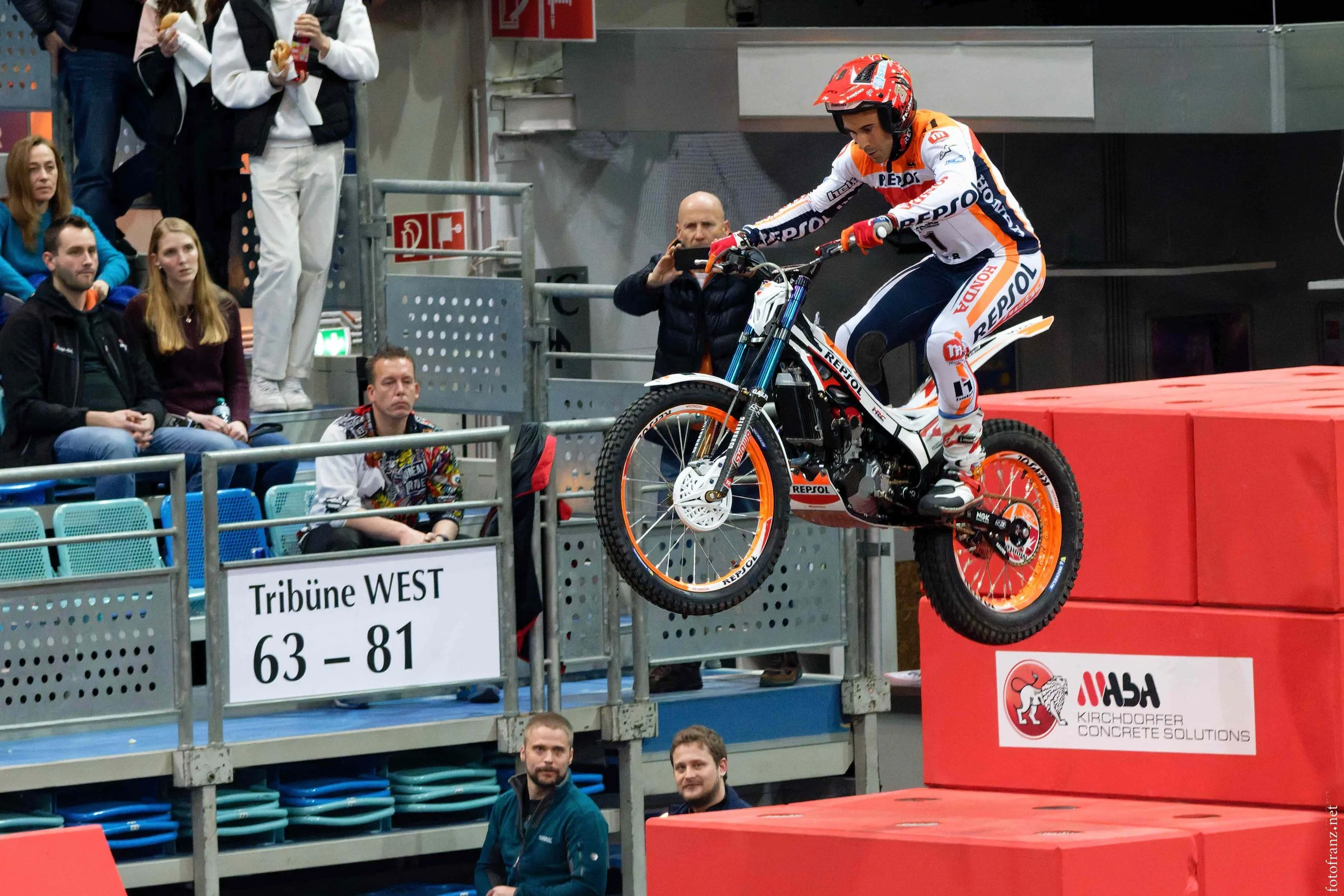 Motorradfahrer bei einem Sprung während eines Wettbewerbs, Zuschauer und Betreuer beobachten. Zeichen für Tribüne 63-81, im Hintergrund rote und graue Wände, Logo des Springer-Wettbewerbs.