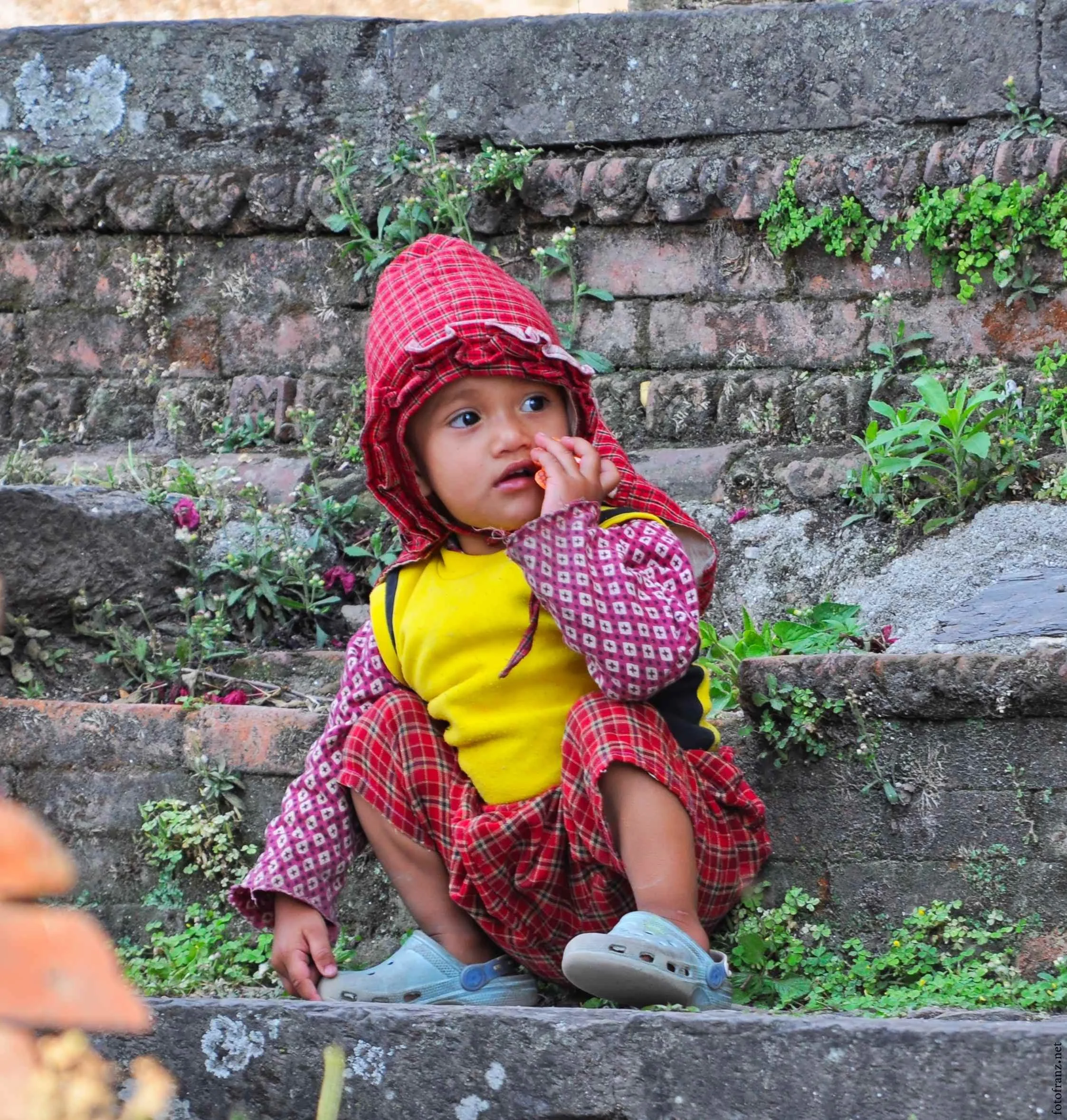 Kleines Kind sitzt auf einer steinernen Treppe vor einer zerbröselten Mauer mit kleinen Pflanzen. Das Kind trägt bunte, kariert gemusterte Kleidung und einen roten, karierten Hut.
