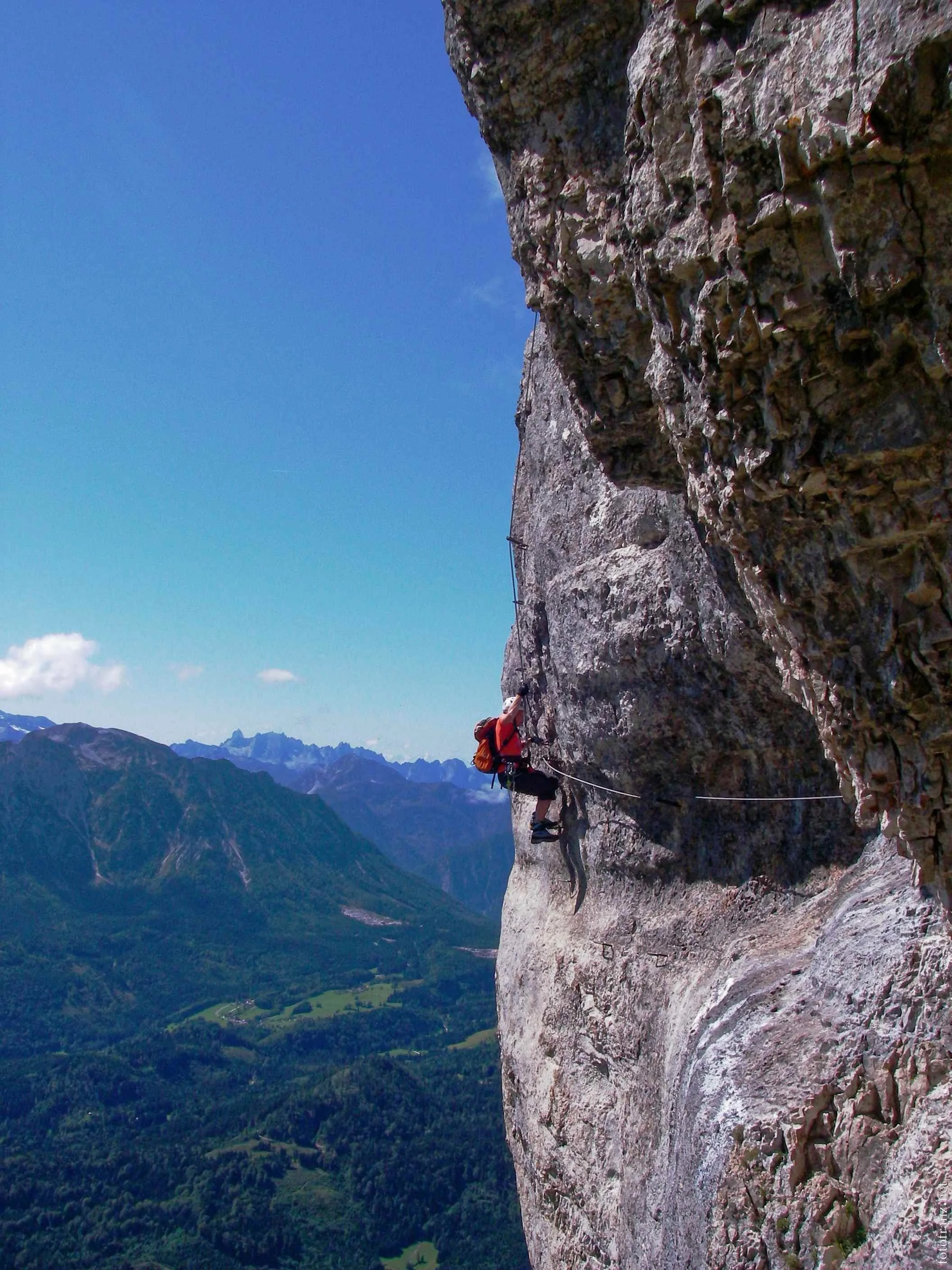 Kletterer erklimmt eine steile Felswand mit Blick auf eine grüne, bergige Landschaft unter blauem Himmel.