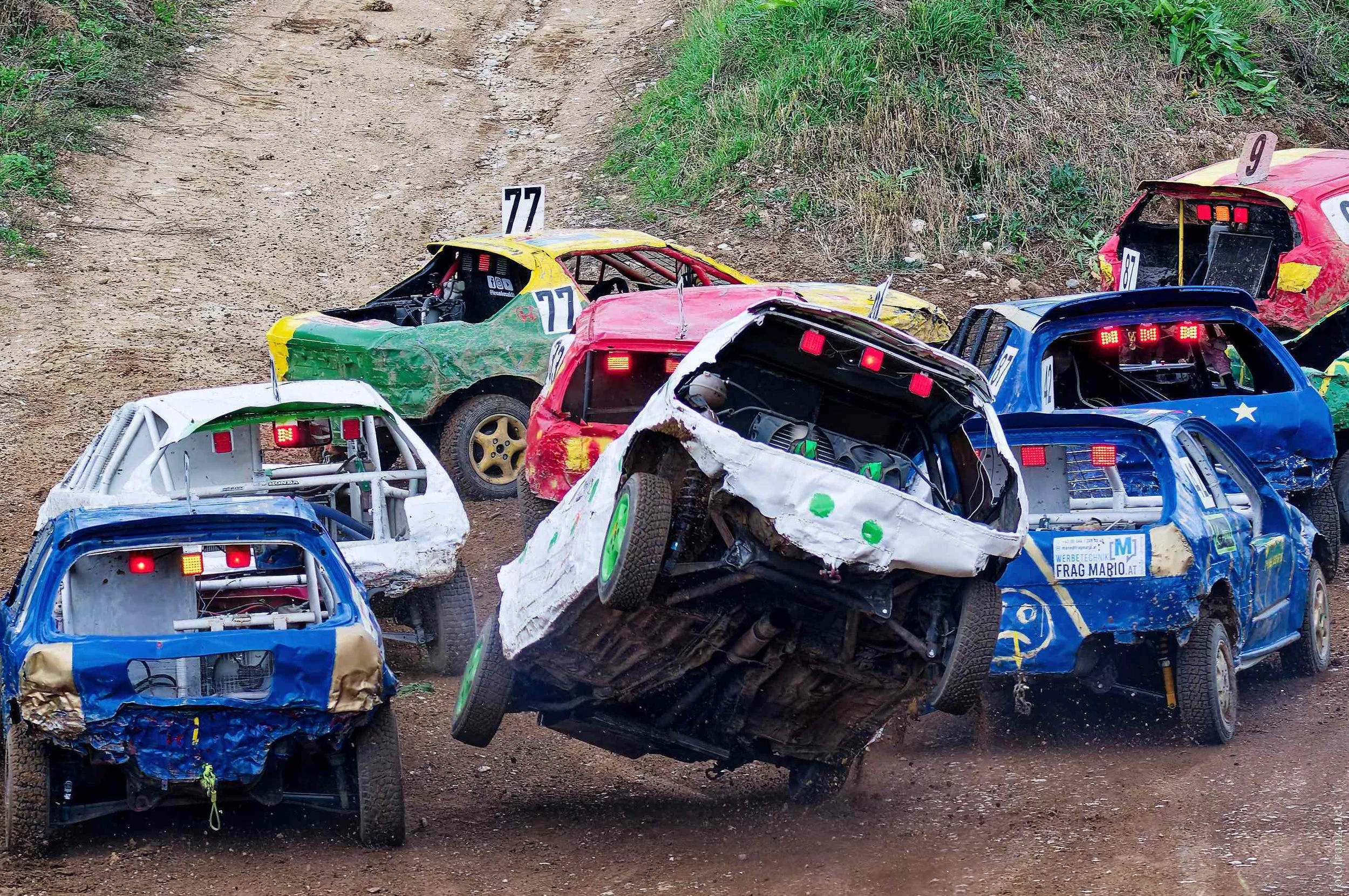 Mehrere zerstörte und beschädigte Rennwagen bei einem Unfall auf einer staubigen Rennstrecke, einige Fahrzeuge sind umgestürzt oder in Bewegung.