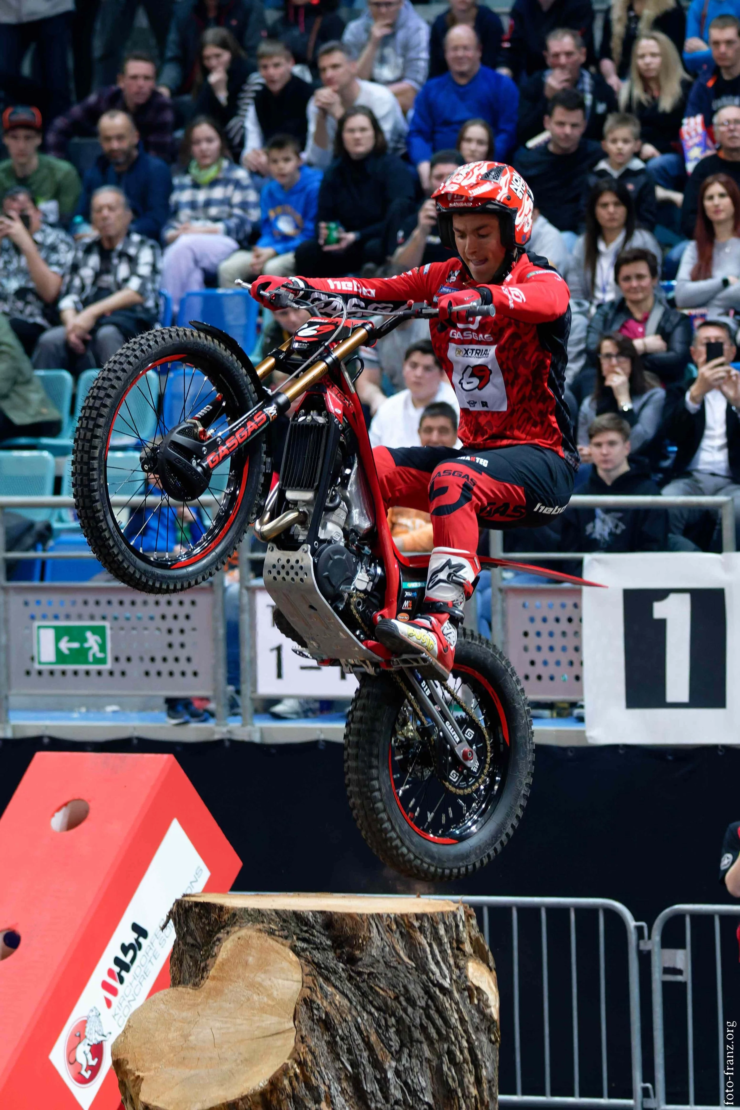 Motorradfahrer springt auf einem Baumstamm bei einer Stunt-Show vor einem Publikum. Das Publikum sitzt auf Tribünen und beobachtet die Aktion.
