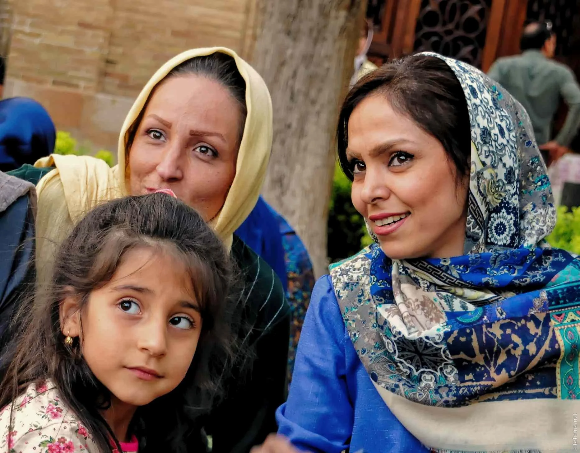 Drei Frauen und ein Mädchen, die nahe beieinander stehen, während sie nach rechts schauen. Alle tragen Kopftücher, und im Hintergrund sind andere Menschen und eine Baum. Die Szene findet im Freien statt.