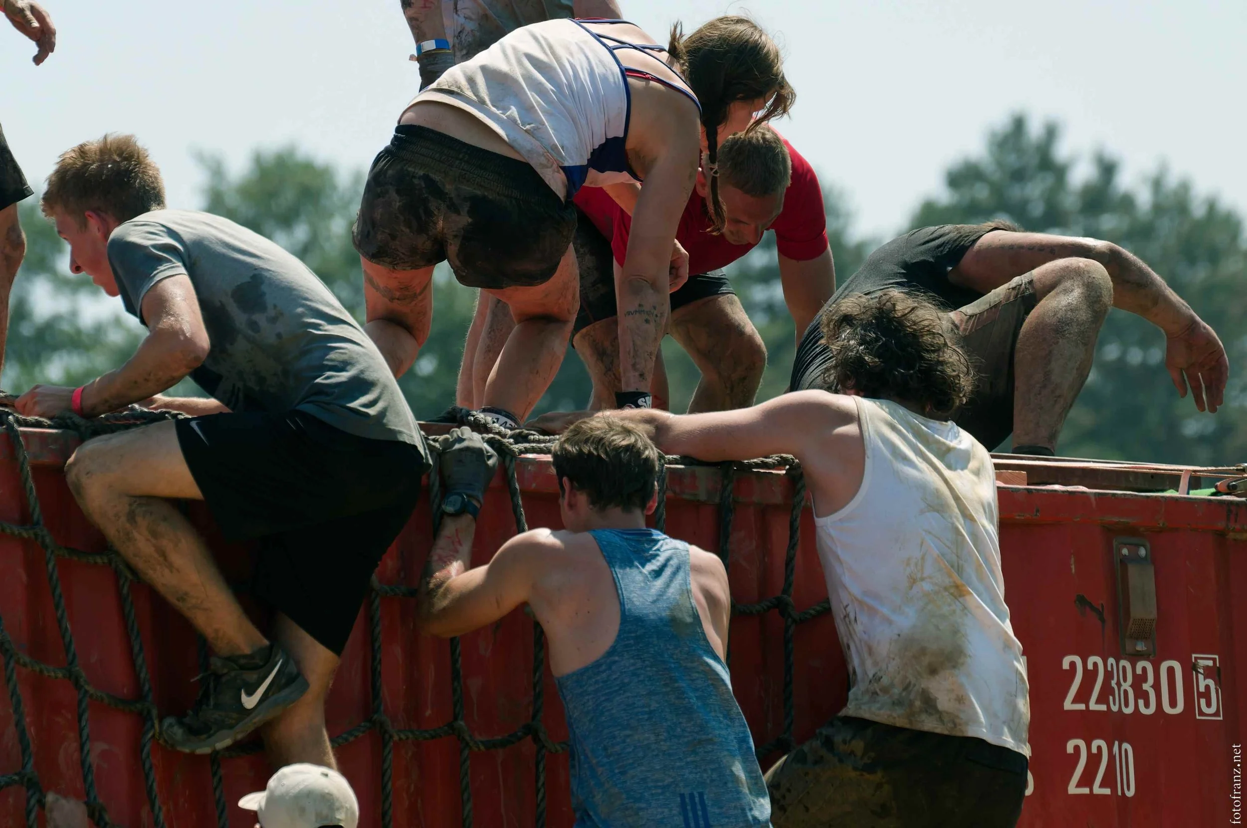 Menschen, die an einem Hindernislauf teilnehmen, steigen auf die Seite eines Containerwagens, sind schmutzig und erschöpft.