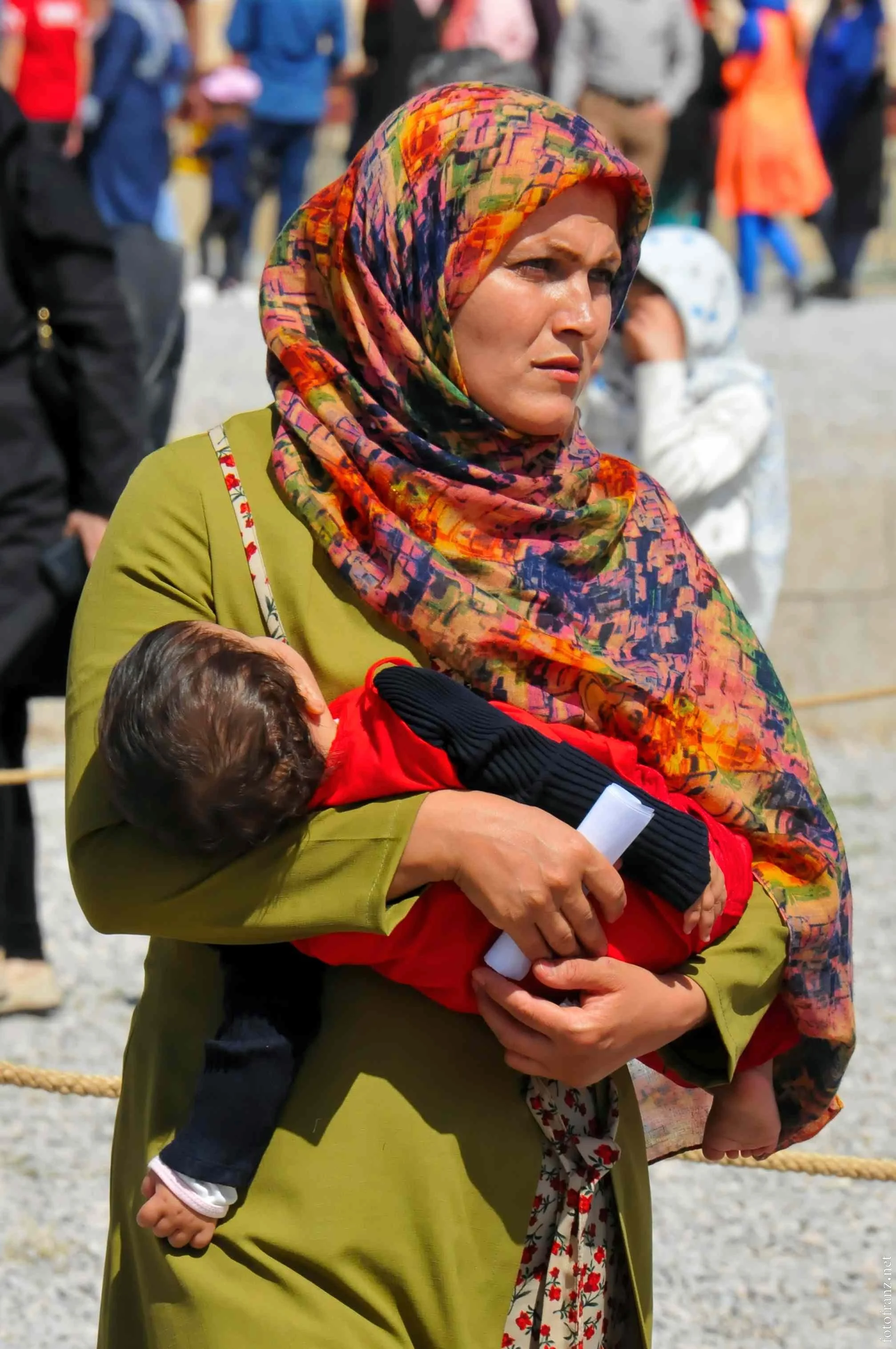 Eine Frau mit einem bunten Kopfscarf hält ein schlafendes Kind in einem roten Jacke in den Armen bei einer Menschenmenge im Hintergrund.