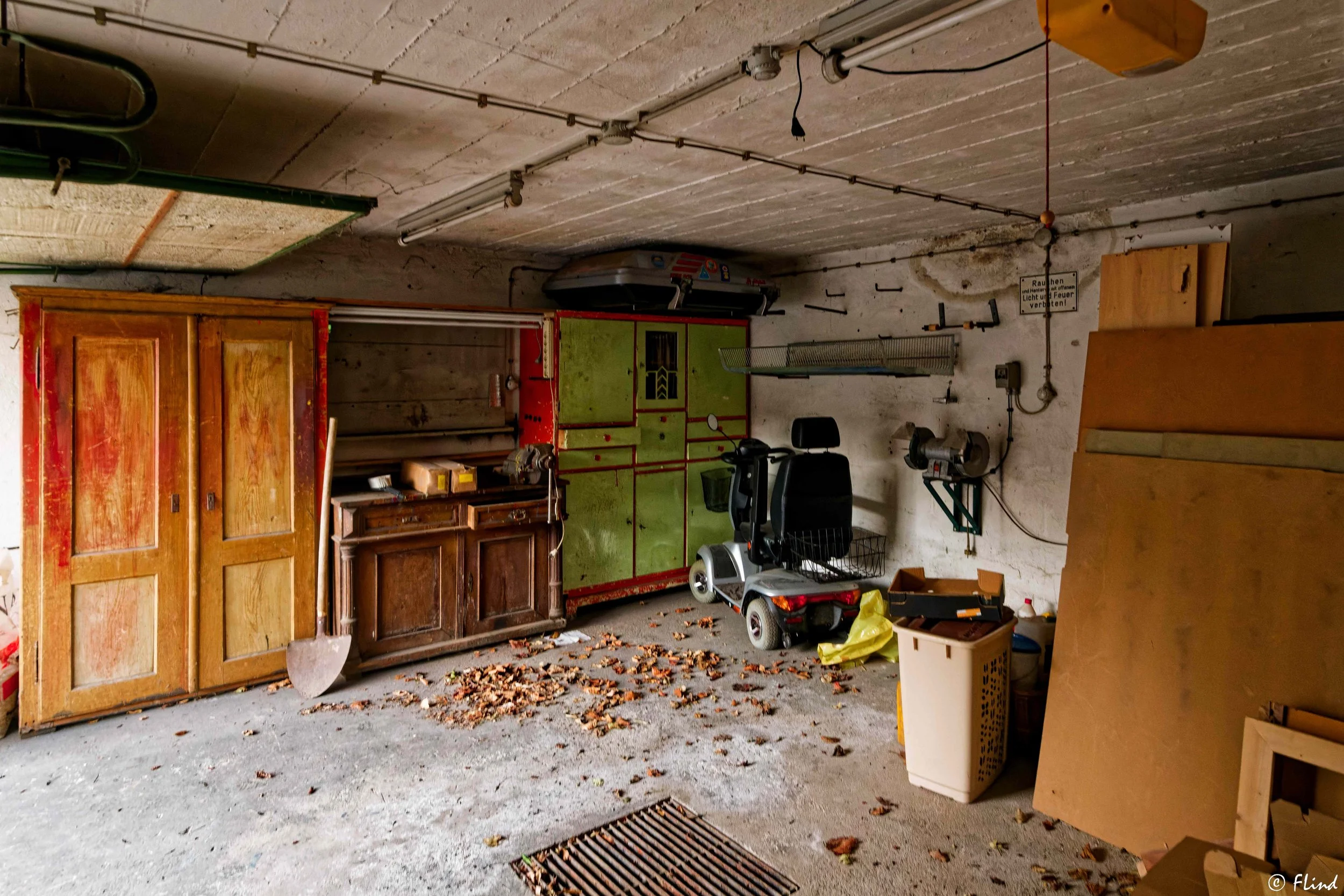 Ein Kellerraum mit alten Möbeln, Werkzeugen, einem elektrischen Rollstuhl, einem grünen Geräteschrank und Kartoffelschalen auf dem Boden, Staub und Blätter liegen verstreut, an der Wand befinden sich Werkzeuge und ein Schild.