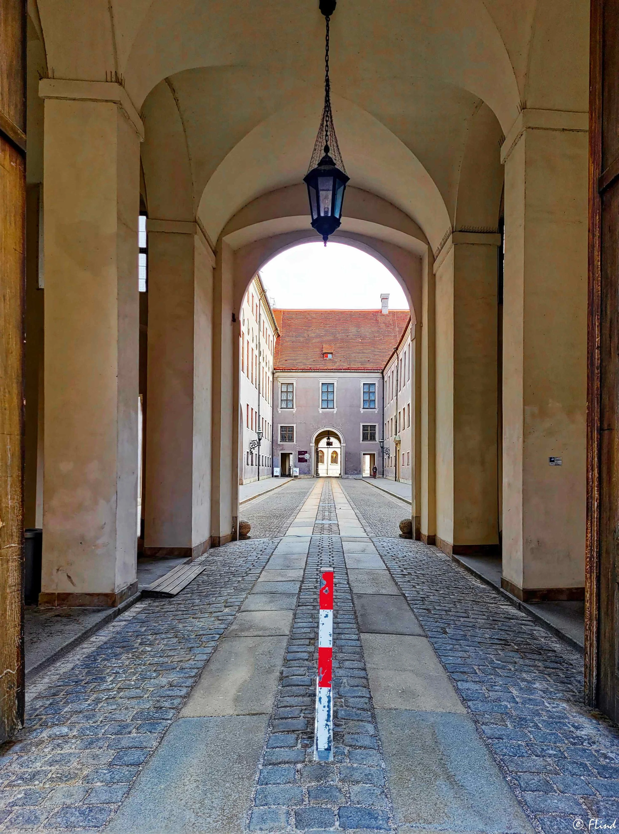 Blick durch einen Torbogen auf einen gepflasterten Innenhof mit mittelalterlichen Gebäuden. Im Vordergrund ist eine rote und weiße Begrenzungssäule auf dem Boden sichtbar. Das Bild ist in der Stadtansicht aufgenommen.