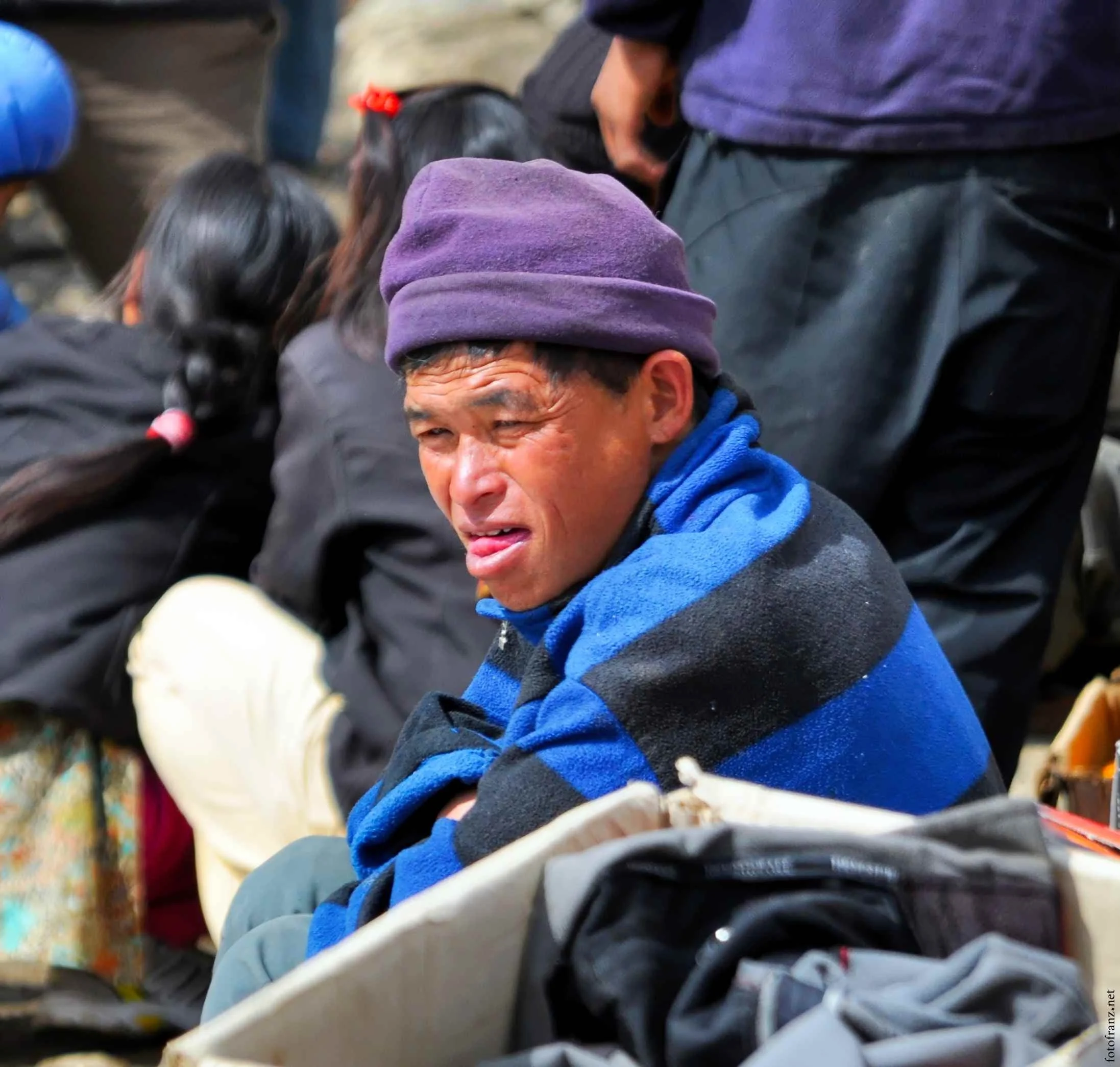 Ein Mann trägt eine lila Mütze und einen blauen Pullover und sieht verzweifelt oder traurig aus. Er sitzt mit anderen Menschen, die auf dem Boden sitzen.